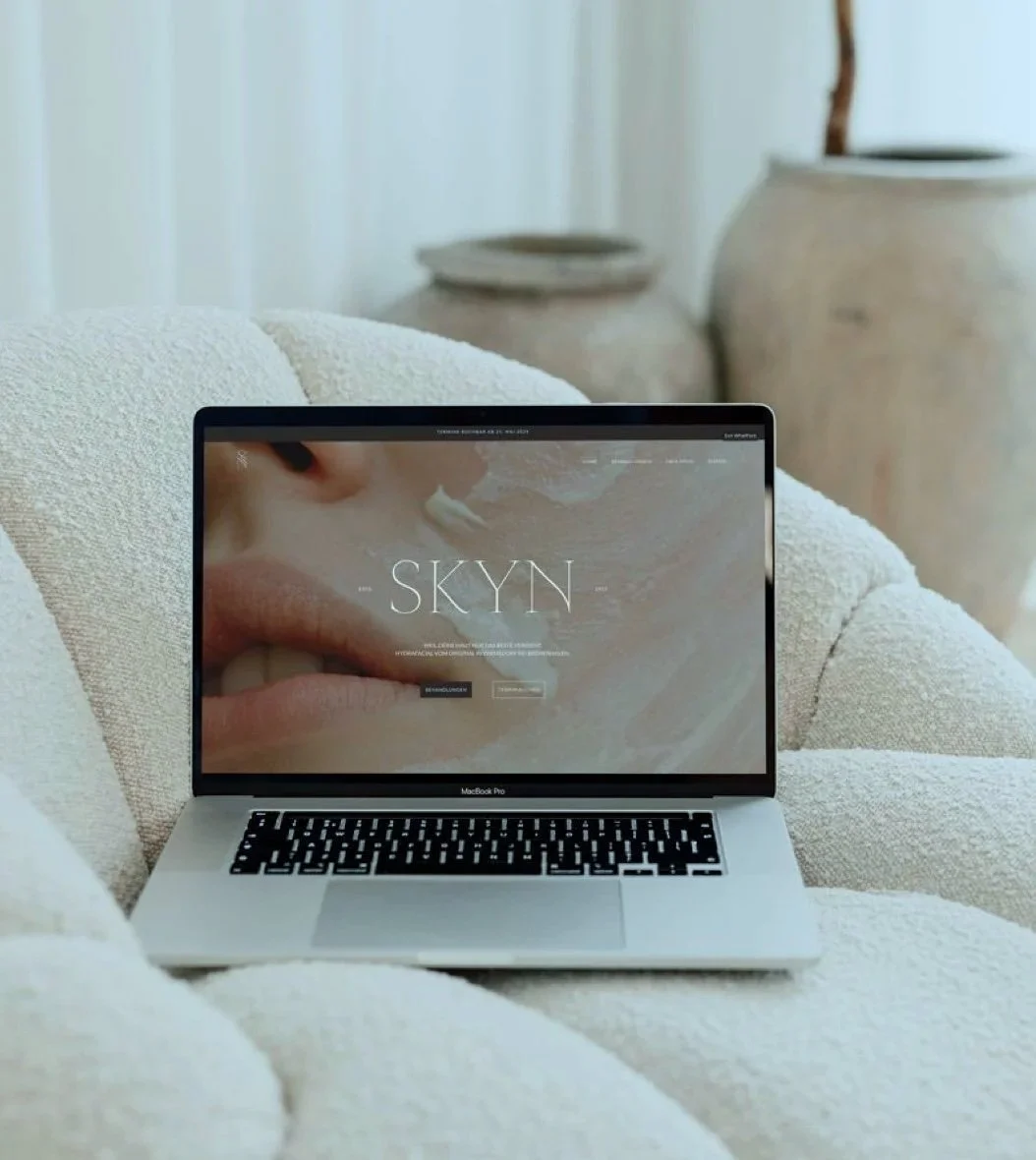 Ein MacBook Pro auf einem weichen, cremefarbenen Sofa mit einem Website-Header, der das Wort 'SKYN' zeigt, und im Hintergrund große alte Keramikvasen.