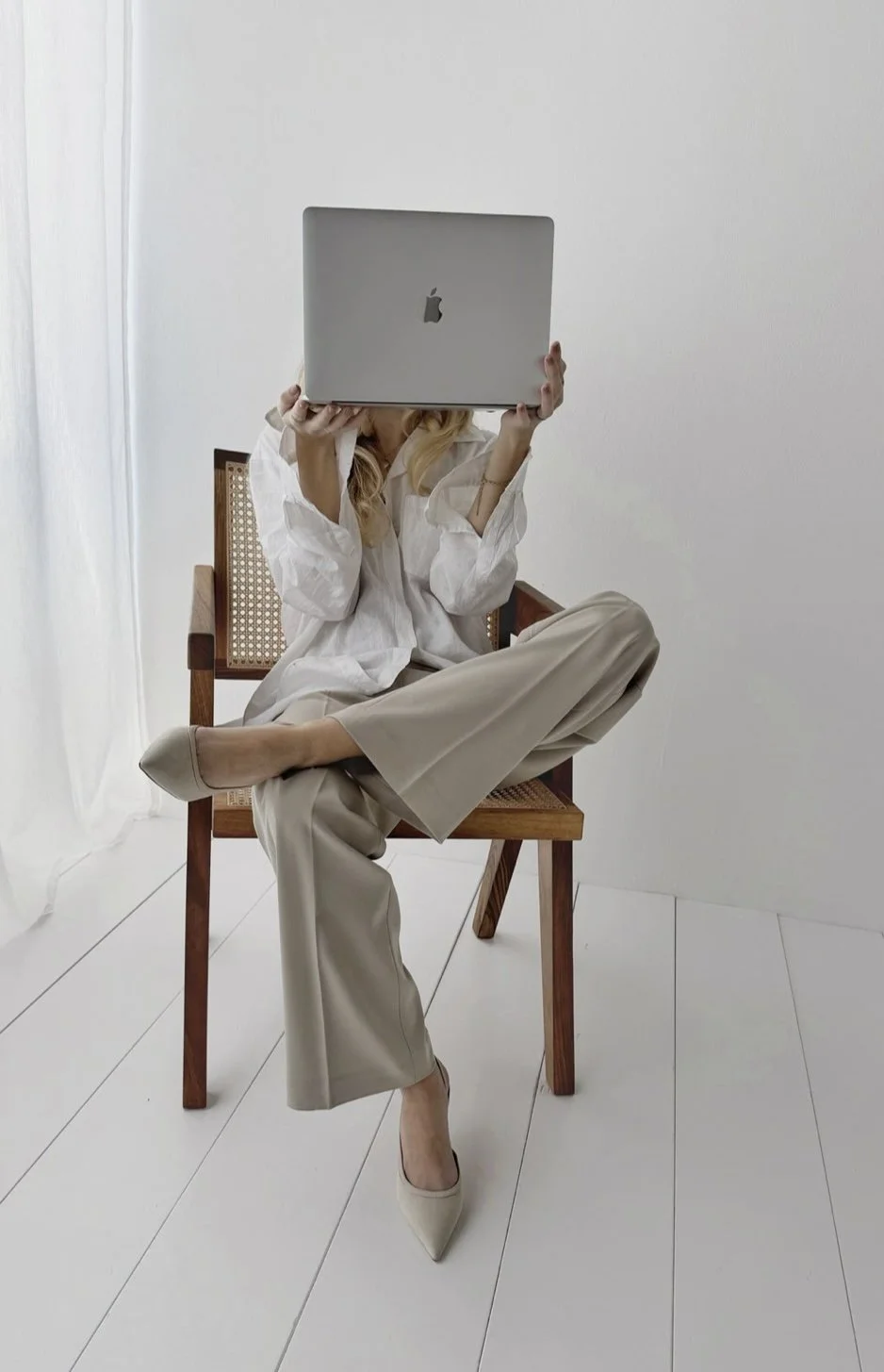 Frau sitzt auf einem Stuhl im weißen Raum, hält einen MacBook-Laptop vor ihr Gesicht, trägt eine weiße Bluse, beige Hosen und weiße High Heels.