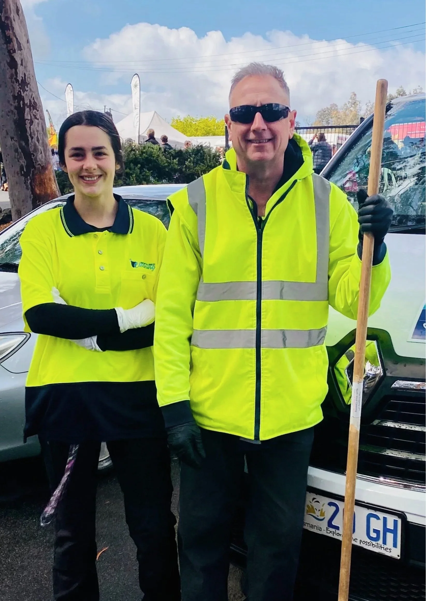 Two event staff members in high-visibility yellow clothing pose beside a car, with one holding a wooden broom handle.