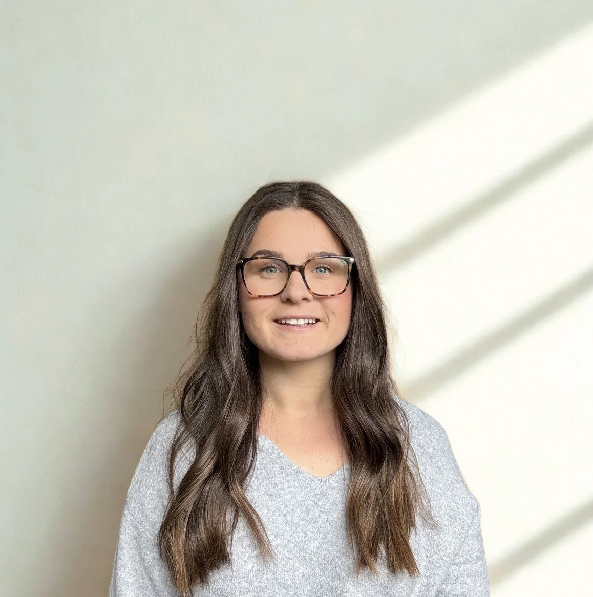 Smiling woman with long brown hair and glasses wearing a gray shirt against a sunlit wall