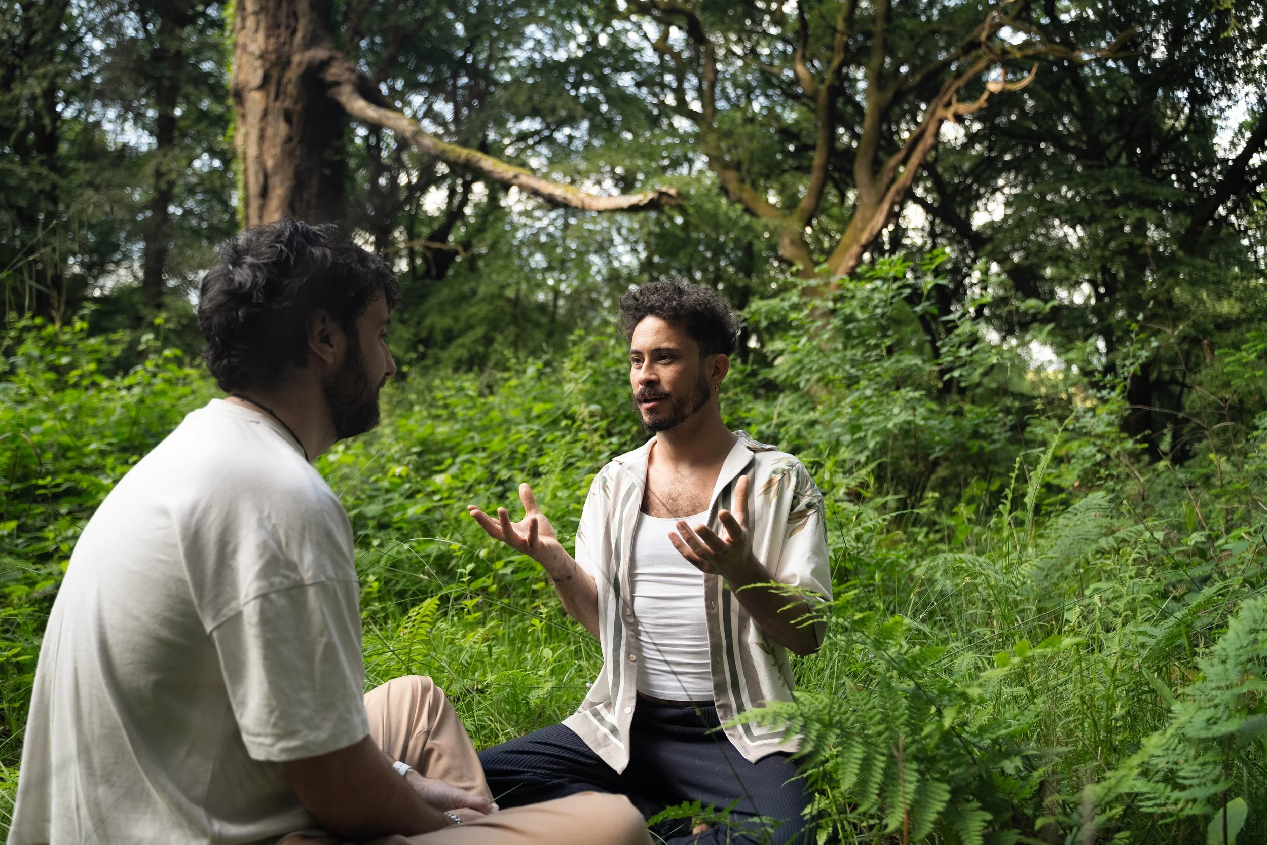 Two men having a conversation in a lush, green forest with dense foliage and tall trees.