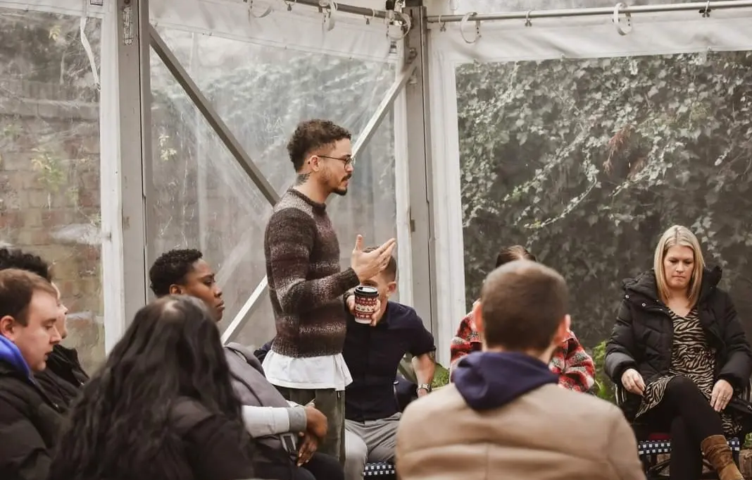 A man in glasses and a striped sweater stands holding a container and speaking to a group of diverse people seated in a semi-circle inside a transparent tent.