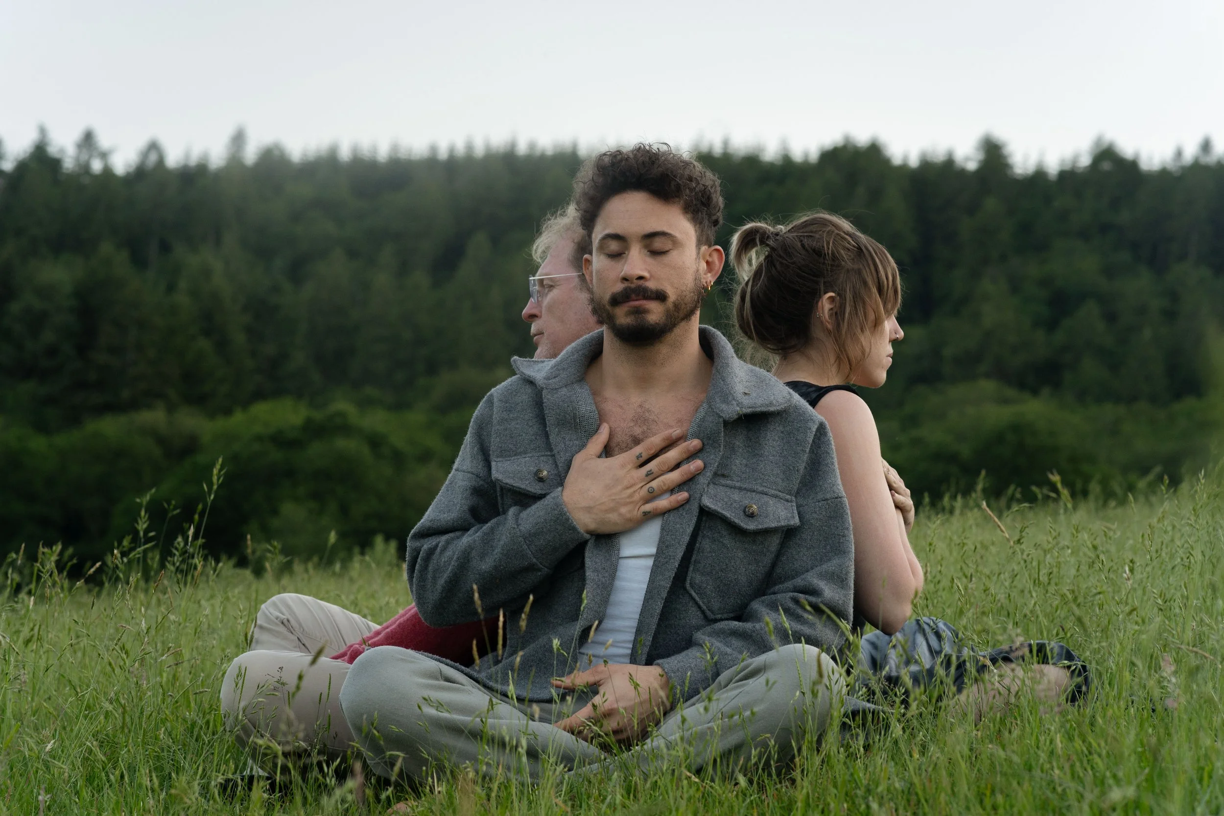 Three people sit on grass in a forest clearing, with their backs to each other, one man with eyes closed and hand on chest, the other two with head in profile, showing diverse appearances.
