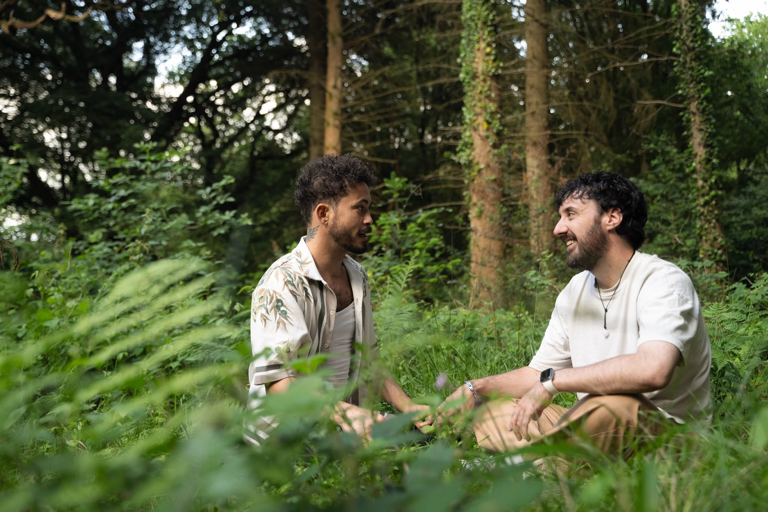 Two men sit on the grass in a lush forest, facing each other and smiling, surrounded by green foliage and tall trees.