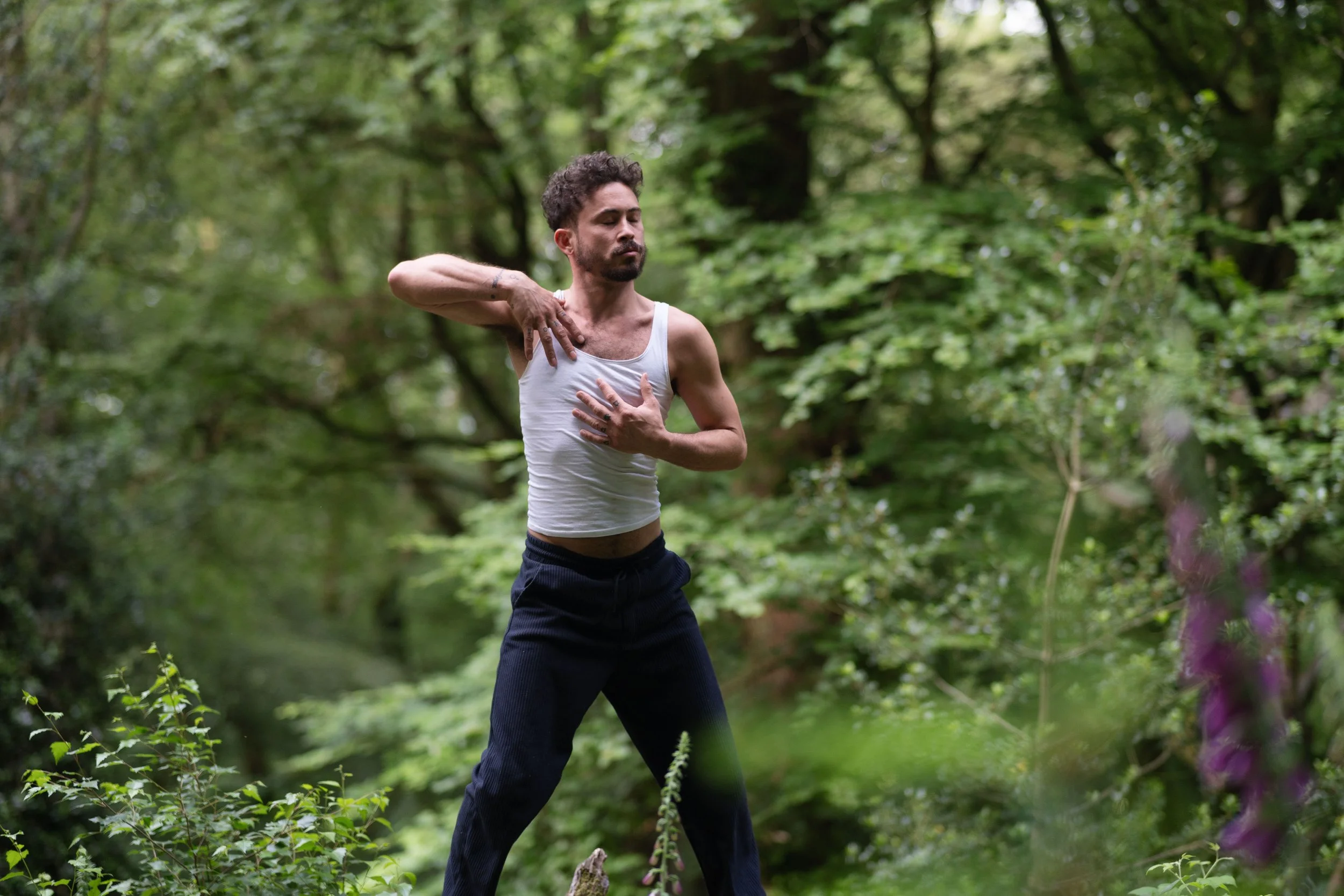 A man with curly hair and tattoos is practicing yoga in a lush green forest. He is wearing a white tank top and black pants, standing with his eyes closed, one hand on his chest and the other on his shoulder, in a meditative pose.