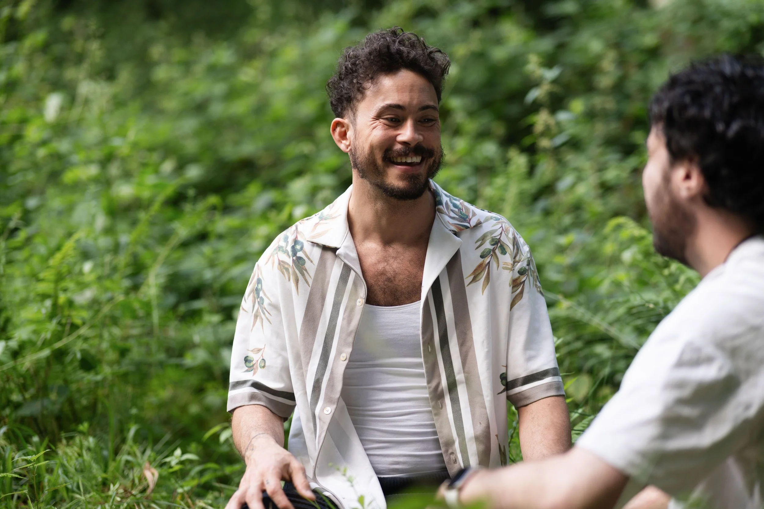 Two men with dark curly hair and beards talking and smiling among green foliage in a lush outdoor setting. One man wears a printed short-sleeve shirt, the other is partially visible.