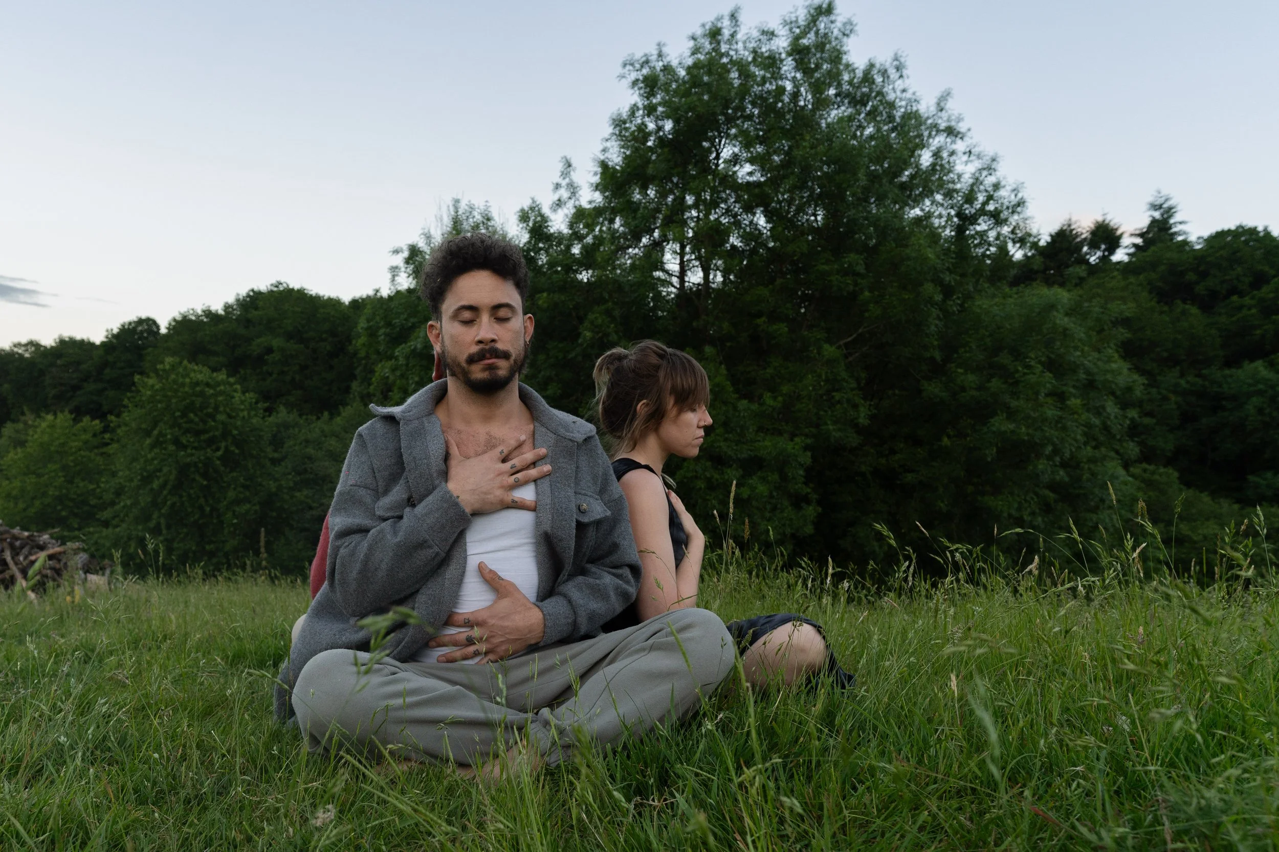 Two people sitting cross-legged in a grassy field, meditating with their eyes closed, surrounded by trees and a clear sky.