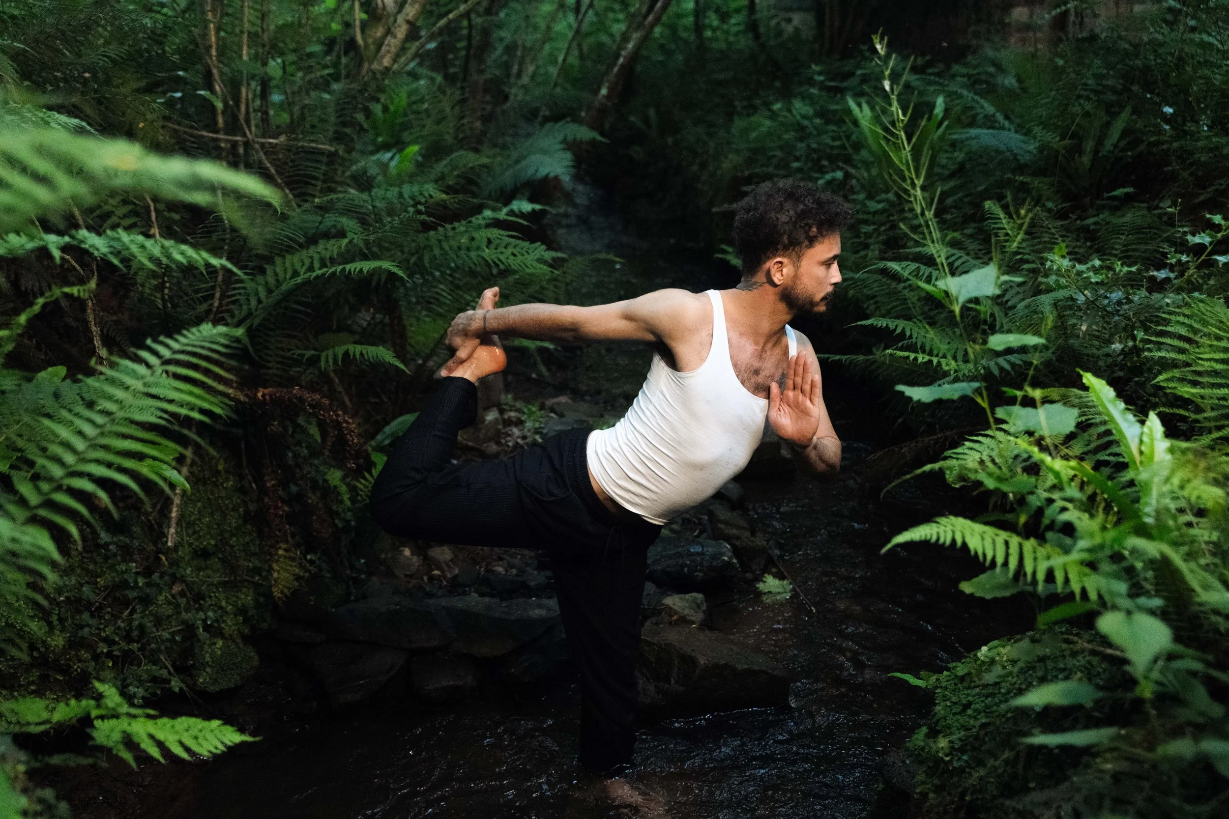 A man practicing yoga outdoors in a lush, green forest, balancing on one leg with one arm extended behind and the other arm in a prayer position in front, near a small stream.