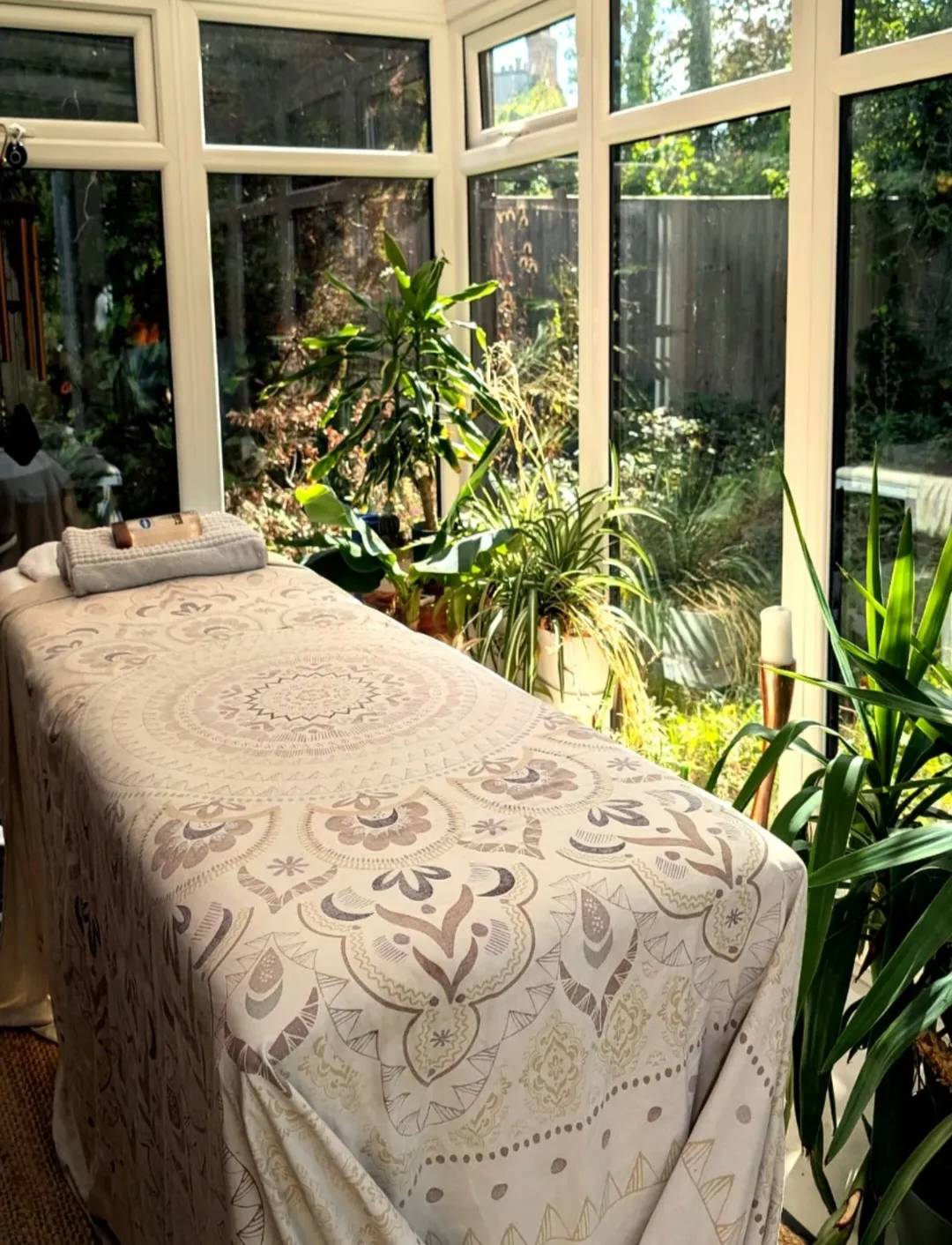 A massage table with a patterned cloth is set up in a glass-enclosed sunroom with large windows, surrounded by green potted plants and sunlight streaming inside.