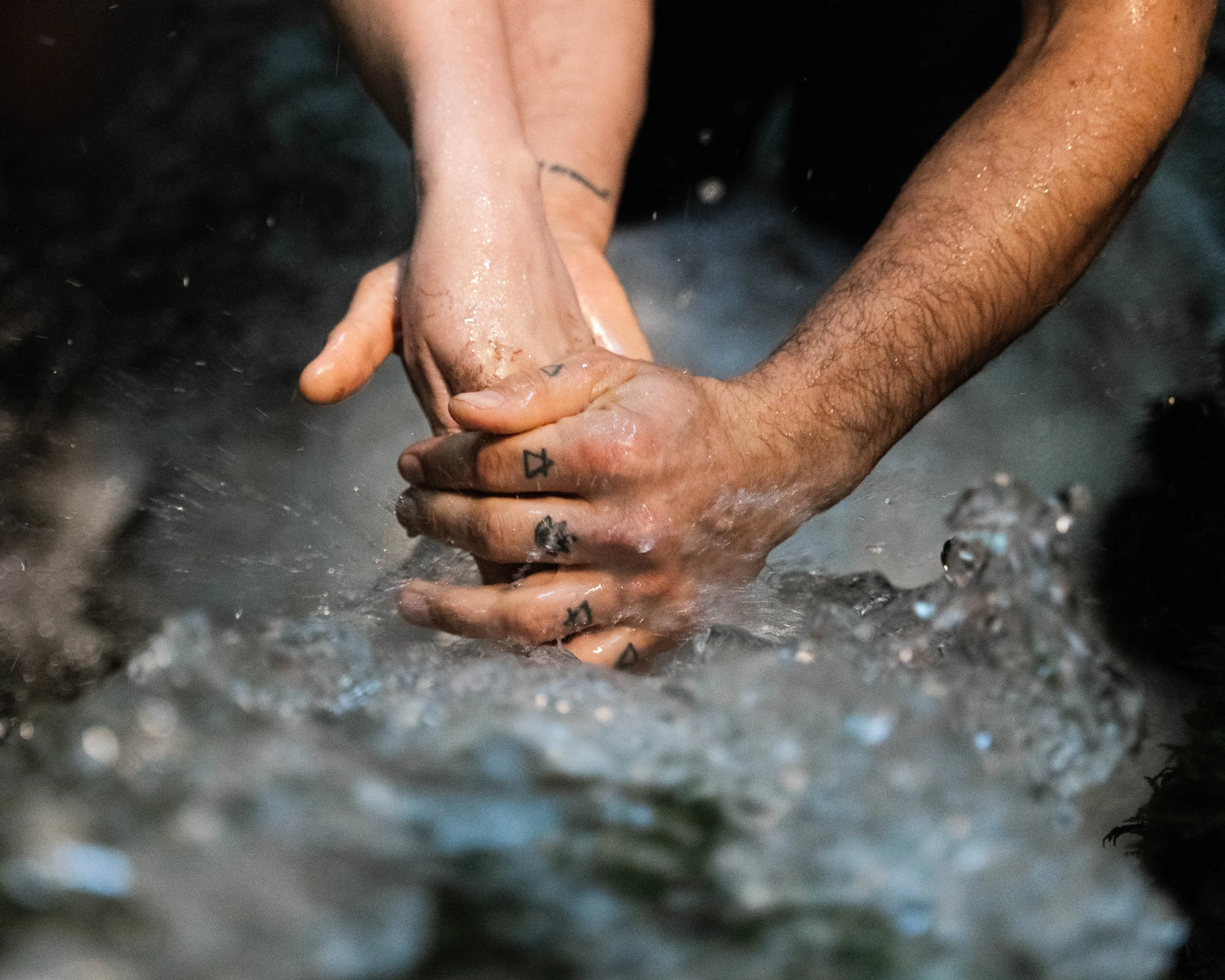 Two hands, one light-skinned and one dark-skinned, clasped together in water, with visible scars and tattoos on the light-skinned hand.
