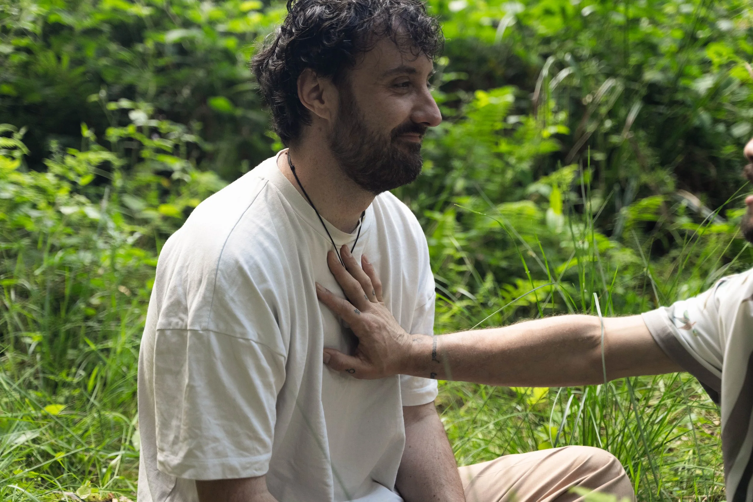 A man with a beard and curly hair sitting in a lush green outdoor setting, with a person touching his chest.