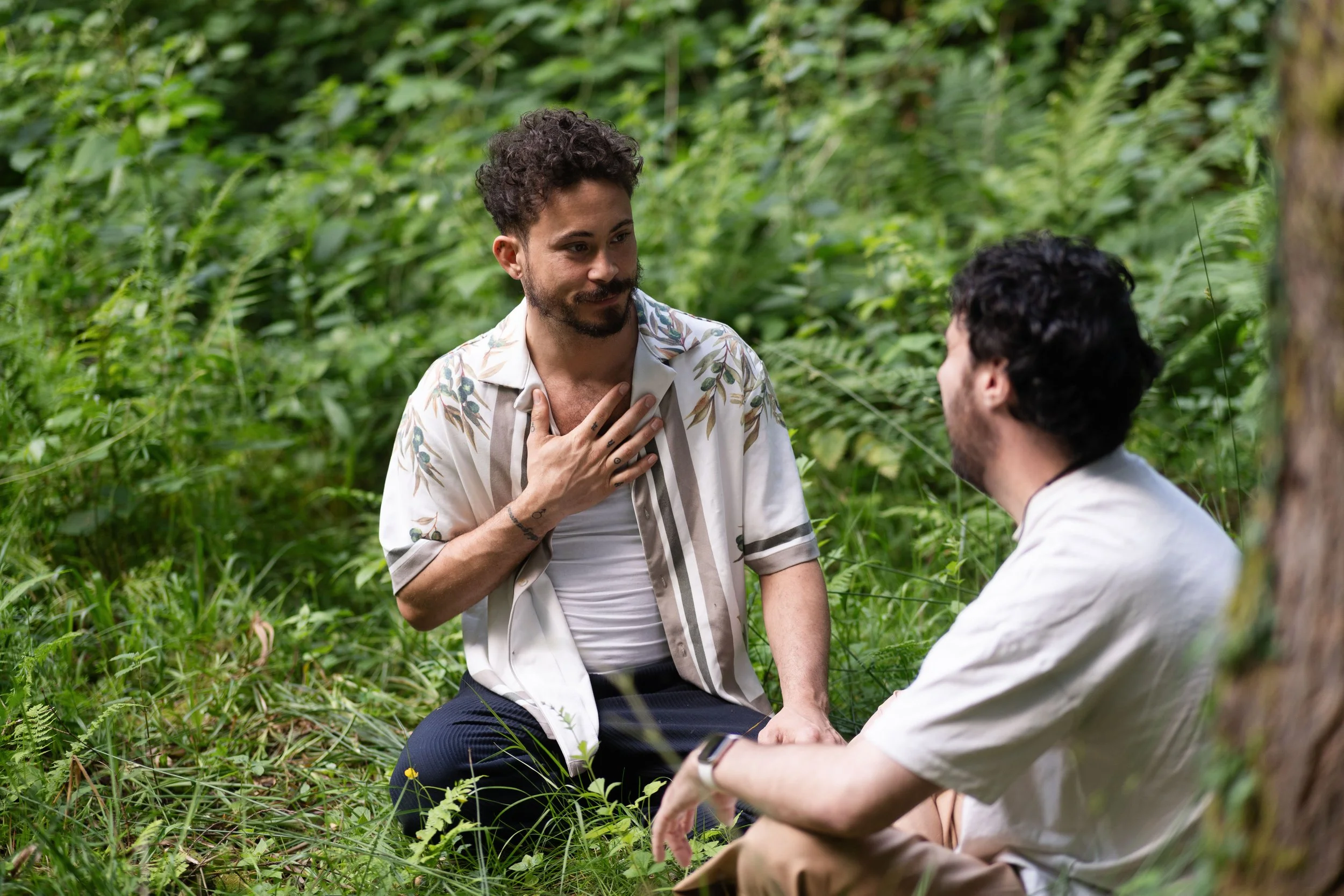 Two men with dark curly hair and beards having a conversation outdoors in a lush green forest. One man is sitting, the other is standing, and the standing man has his hand on his chest.