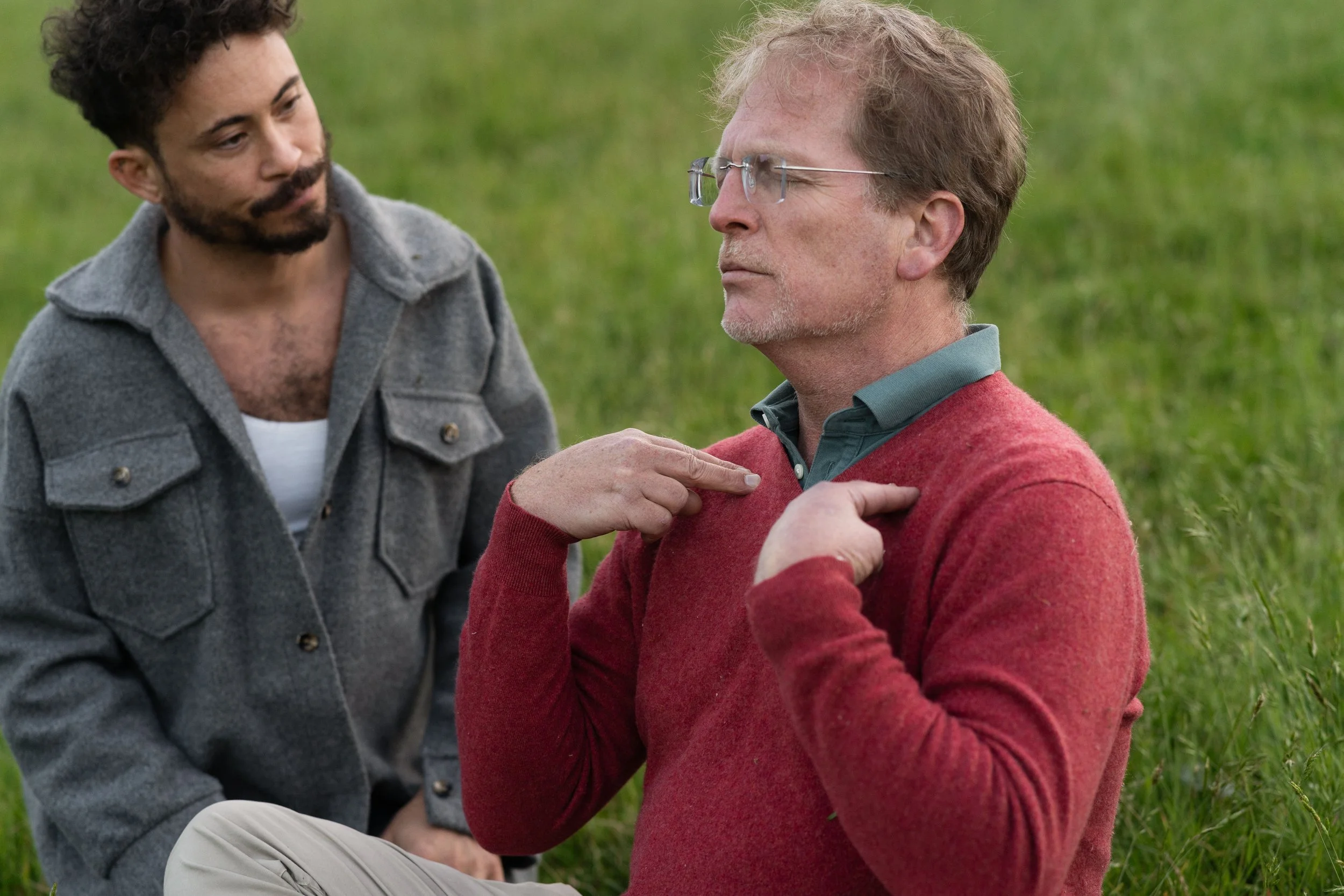 Two men are outdoors, one with gray hair and glasses wearing a red sweater, and the other with dark curly hair and a beard wearing a gray jacket. The man in red is touching his chest while looking solemn, and the other man is looking at him with concern, with green grass in the background.