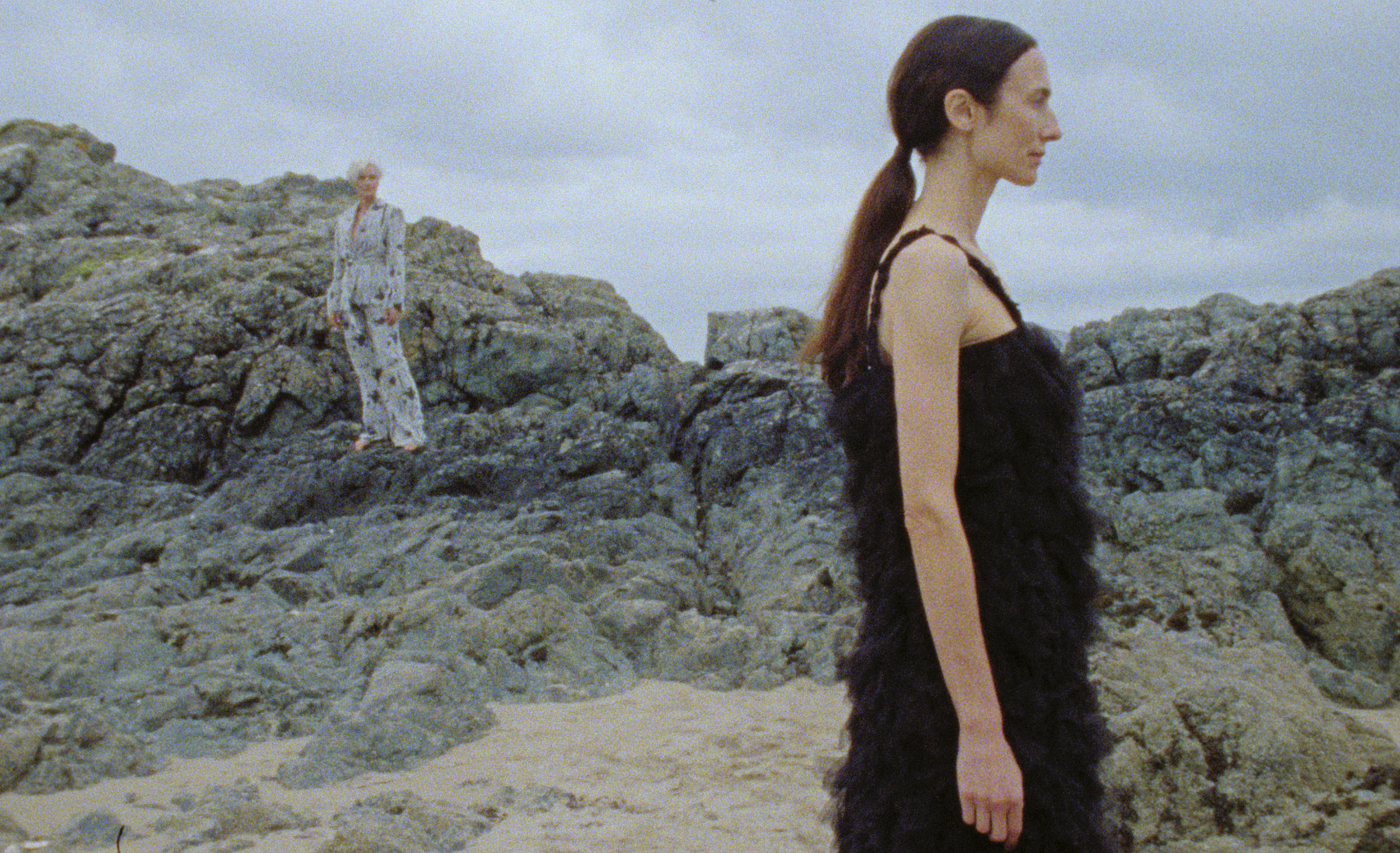 Woman with long dark hair tied back, wearing a black dress with lace details, standing on a rocky beach, facing sideways.