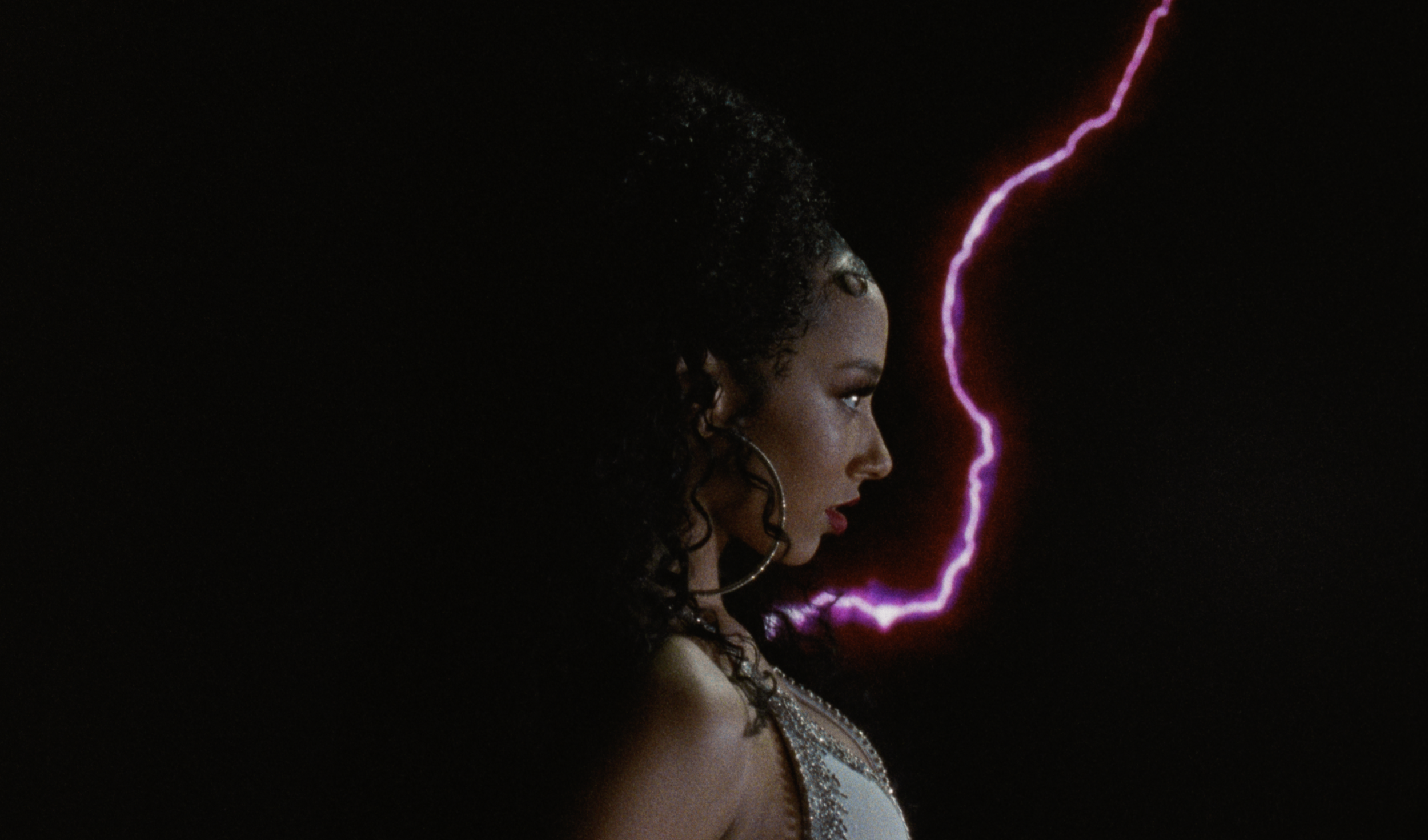 A side profile of a woman with dark, curly hair, wearing hoop earrings, standing against a dark background with a purple lightning bolt.