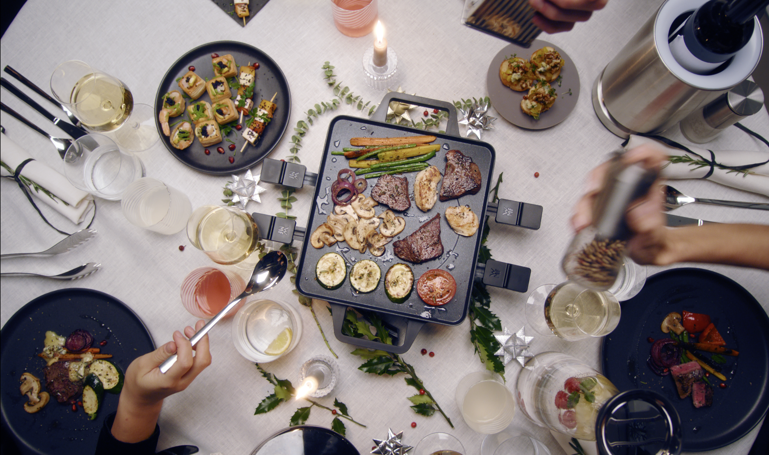 Top-down view of a festive table with various dishes, glasses of white wine, candles, and decorative elements. There is a grill with cooked vegetables and meats, and sushi on plates, with utensils and napkins arranged around.