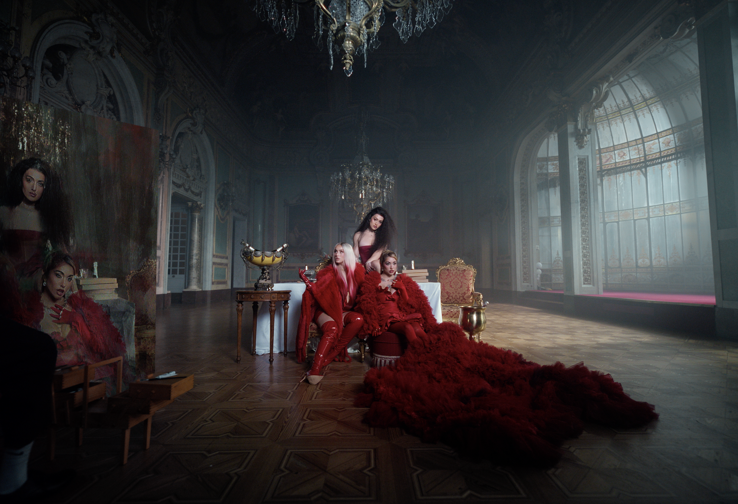 An opulent room with ornate chandeliers, tall windows with stained glass, and decorative molding. Four women dressed in red outfits with feathers and thigh-high boots pose on and around a white sofa, with one woman sitting on a chair nearby. There is