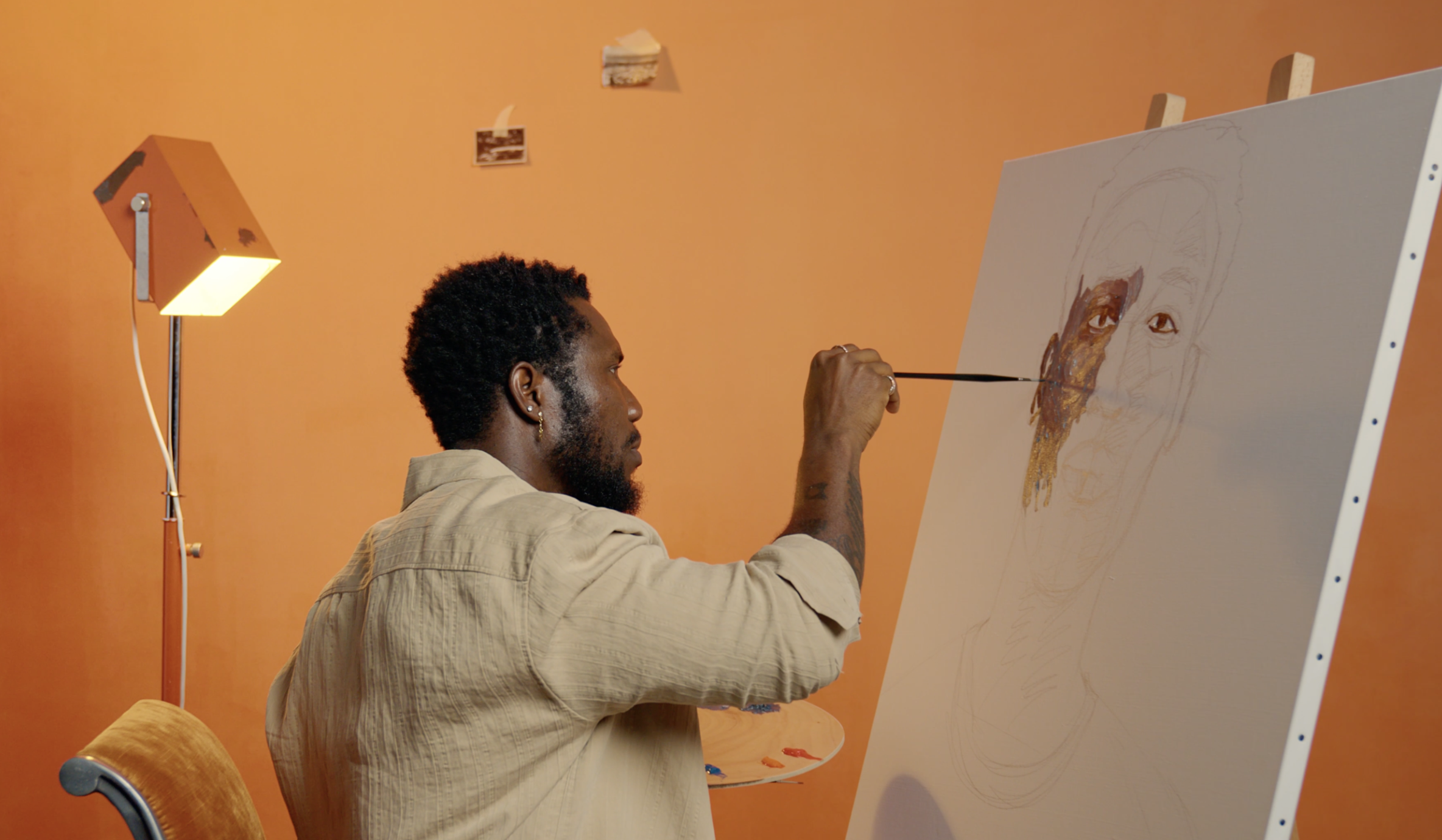 An artist with curly black hair, beard, and earrings paints a portrait of a face on a large canvas with a palette. The room has orange walls and a warm, expressive atmosphere.