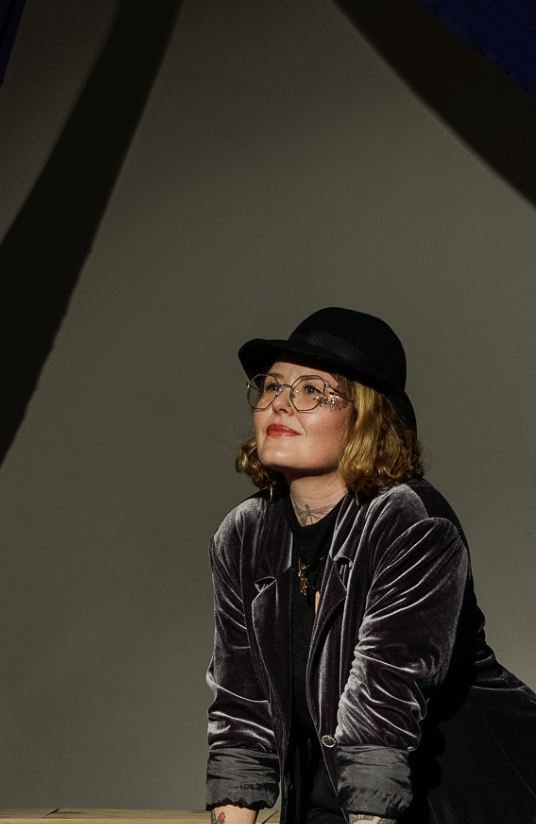 A woman with glasses and shoulder-length curly hair, wearing a black hat and velvet jacket, sitting at a table with a neutral background.