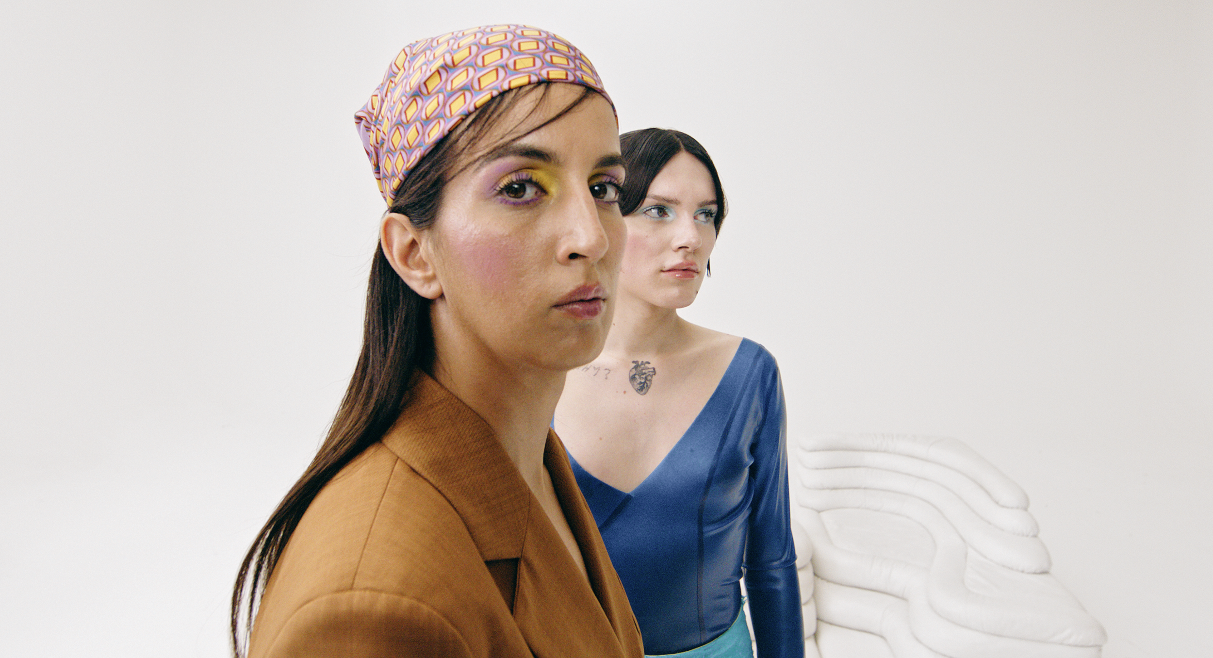 Two women stand against a white background with abstract white sculpture. The woman in front has long brown hair, wearing a patterned headscarf and a brown blazer. The woman behind has black hair, wearing a blue dress, with a tattoo on her chest.