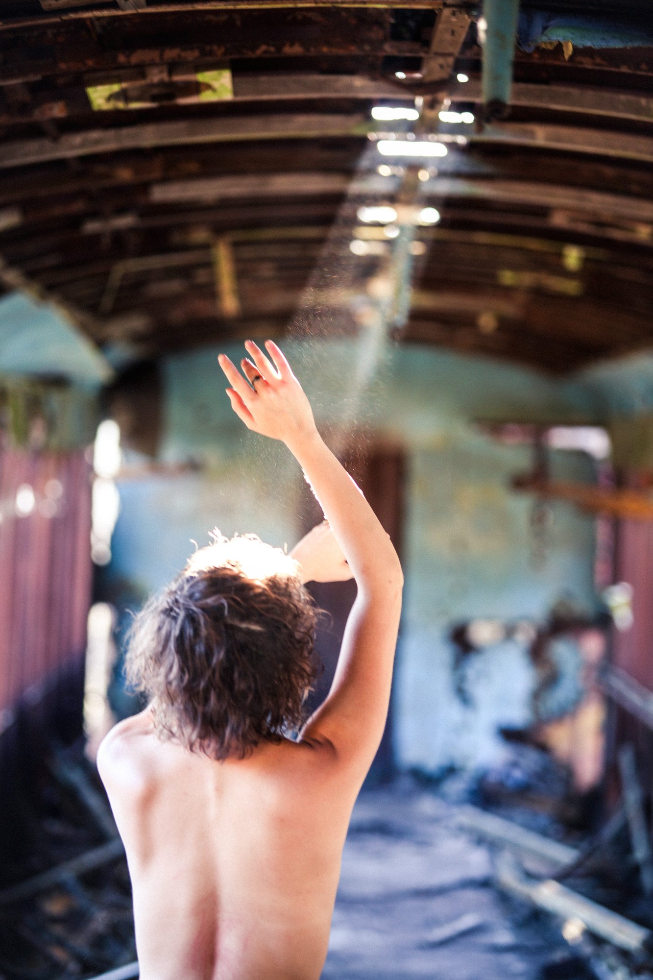 Young person with short curly hair, no shirt, reaching up with their right hand in motion, in a rustic setting with wooden beams and blurred background.