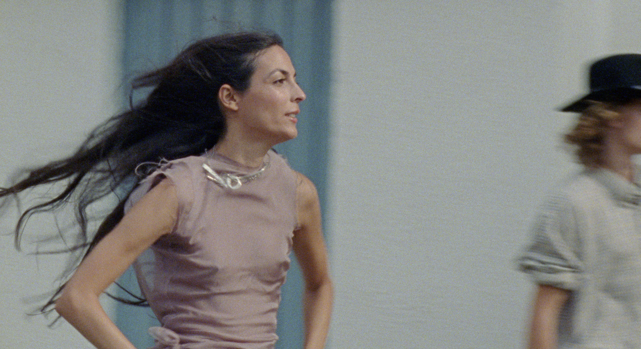 A woman with long dark hair running in an indoor space, wearing a light purple top with a metallic necklace.