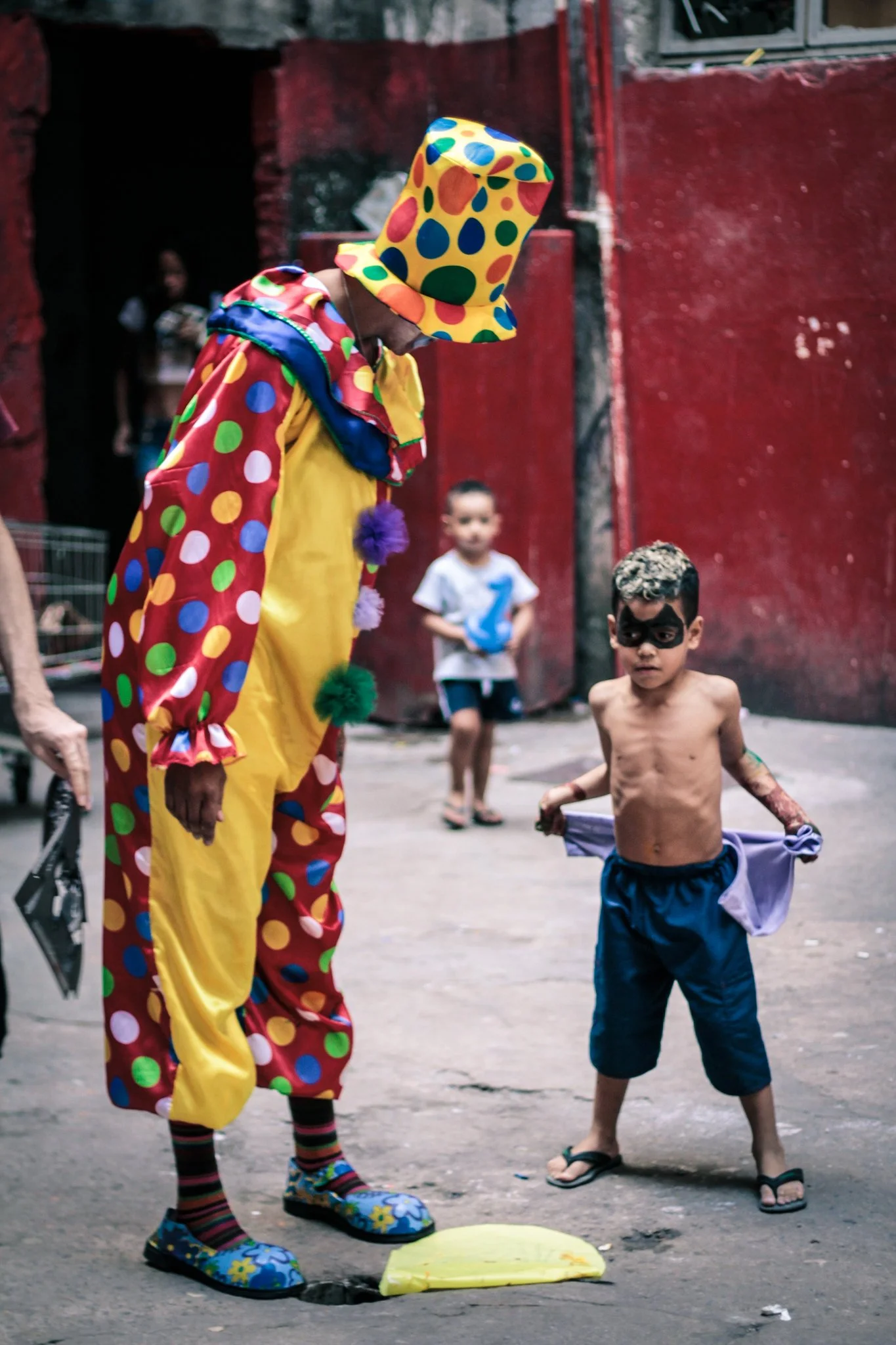 A person dressed as a clown in colorful polka dot costume and hat standing near a small, shirtless child wearing face paint, in an outdoor urban setting with a red wall in the background. The clown appears to be observing or interacting with the chil