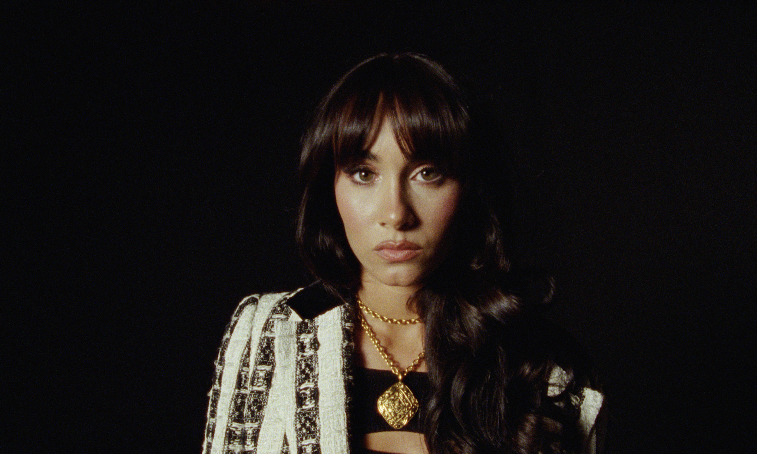 A woman with dark hair and brown eyes wearing a black top, gold jewelry, and a patterned cream and black jacket, against a black background.