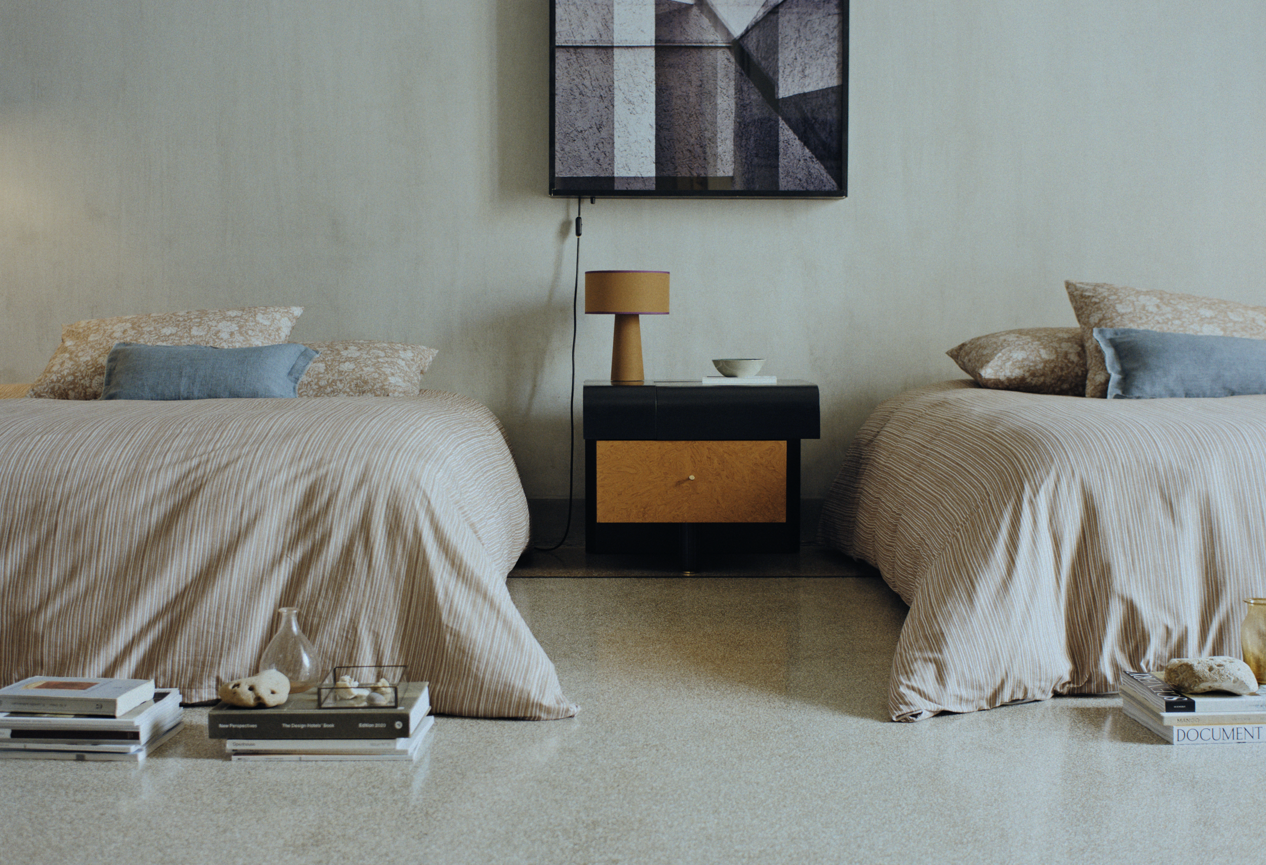 Twin beds in a bedroom, each with beige and blue bedding and pillows. A black nightstand with a mustard-colored lamp and other items separates the beds. A framed abstract artwork hangs above the nightstand. Items like books, cookies, and a remote are