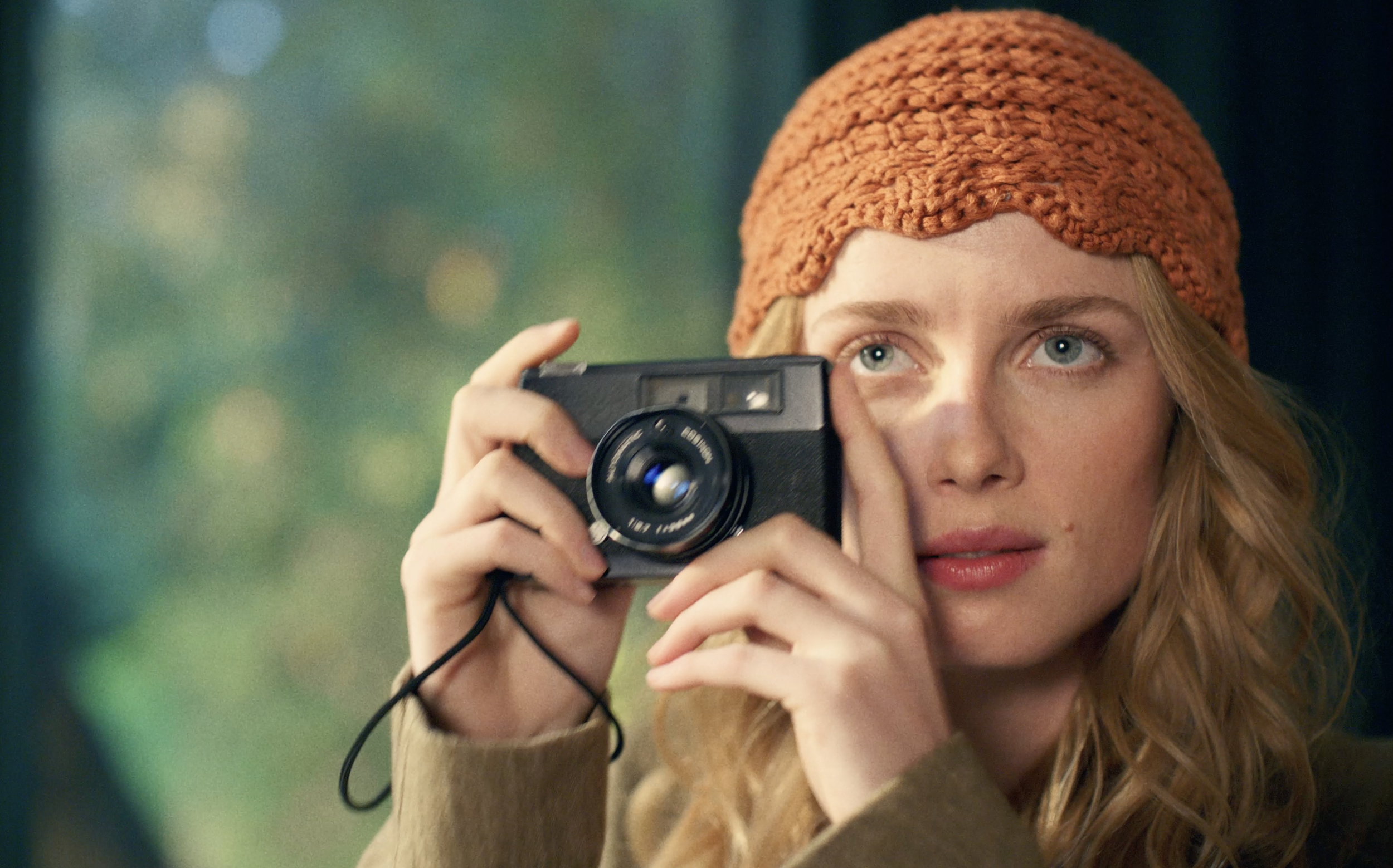 A young woman with curly hair wearing a knit orange hat and a beige coat, taking a photo with a black camera. She has blue eyes and is outdoors with blurred greenery in the background.