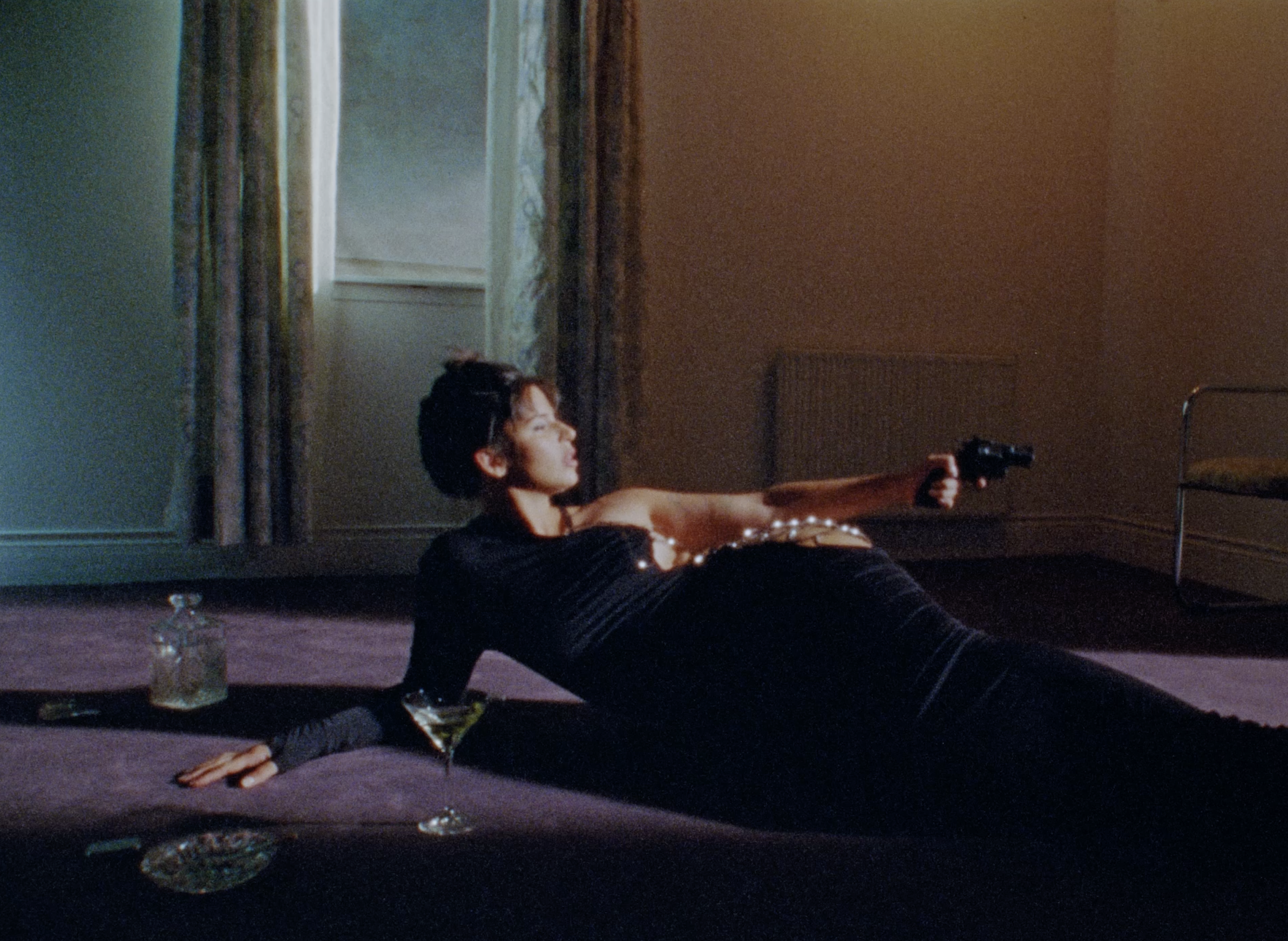 A person lying on a couch in a room, holding a gun, with a table and glasses nearby, in a dimly lit space.