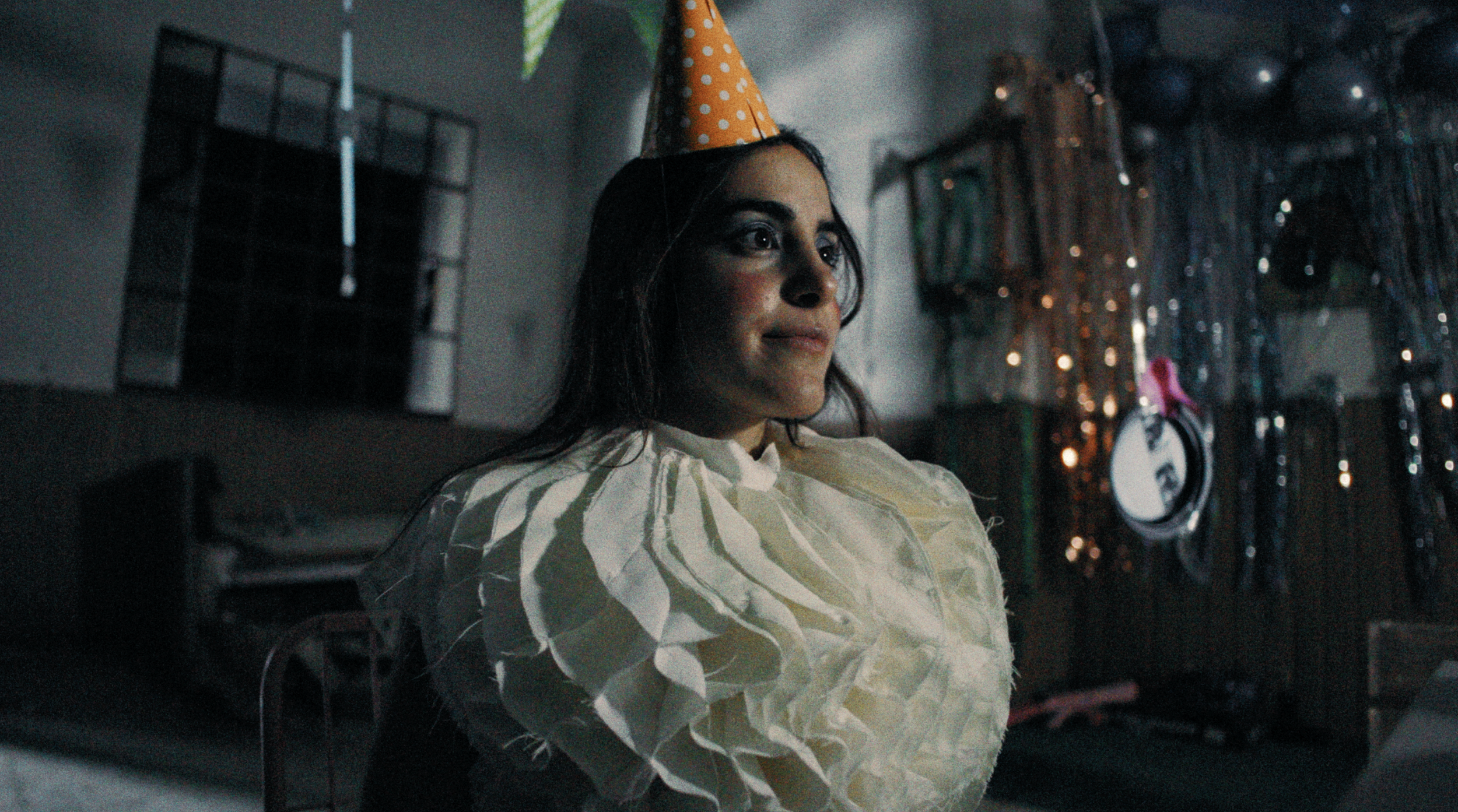 A woman with long dark hair wearing an orange party hat with white polka dots and a cream-colored robe, sitting in a dimly lit room decorated with balloons and metallic streamers, looking thoughtfully to the side.