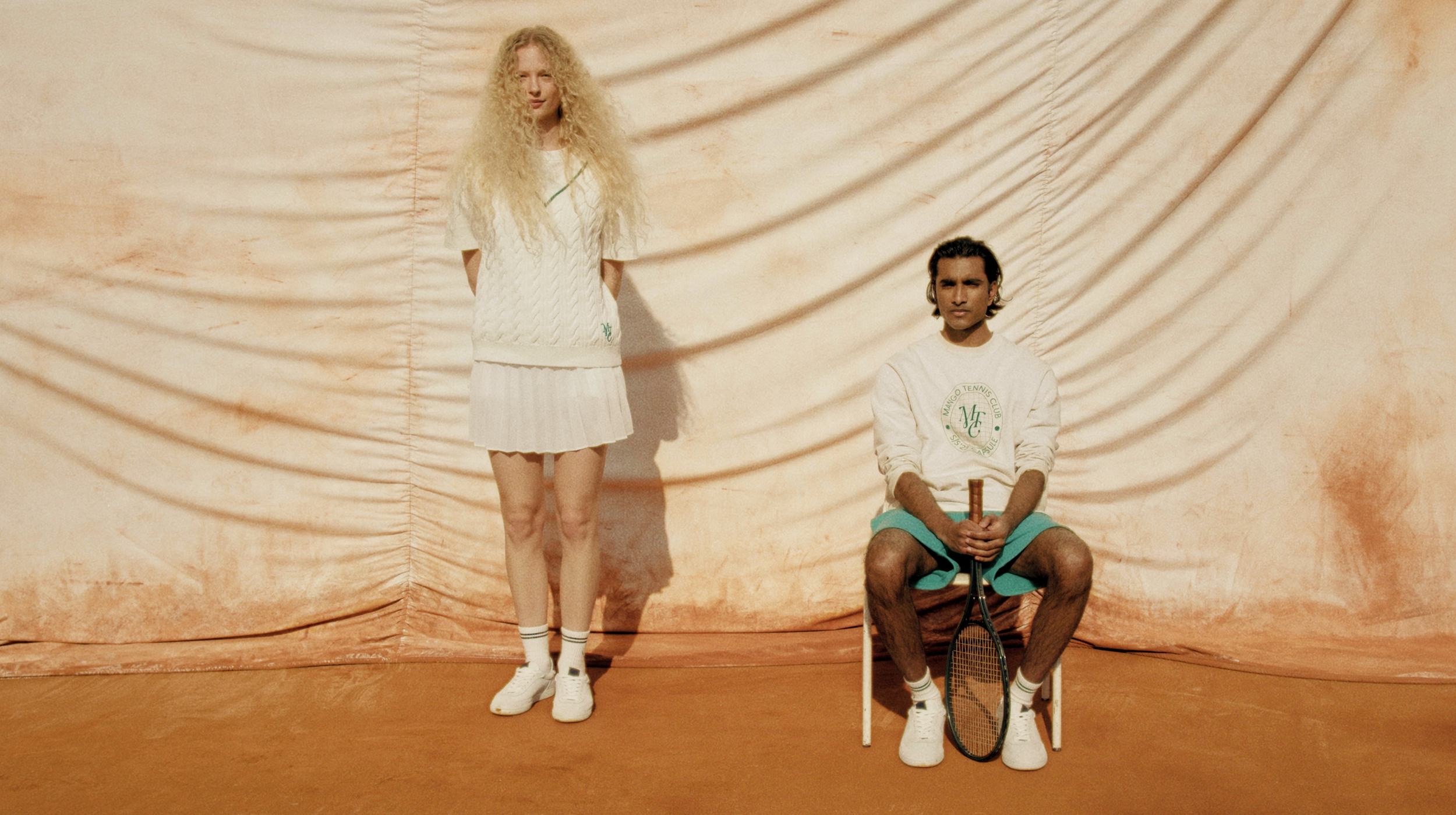 A woman with long curly blonde hair standing with her hands behind her back and a man with dark hair sitting on a chair holding a tennis racket, both dressed in tennis clothing, on a tennis court with a beige backdrop.