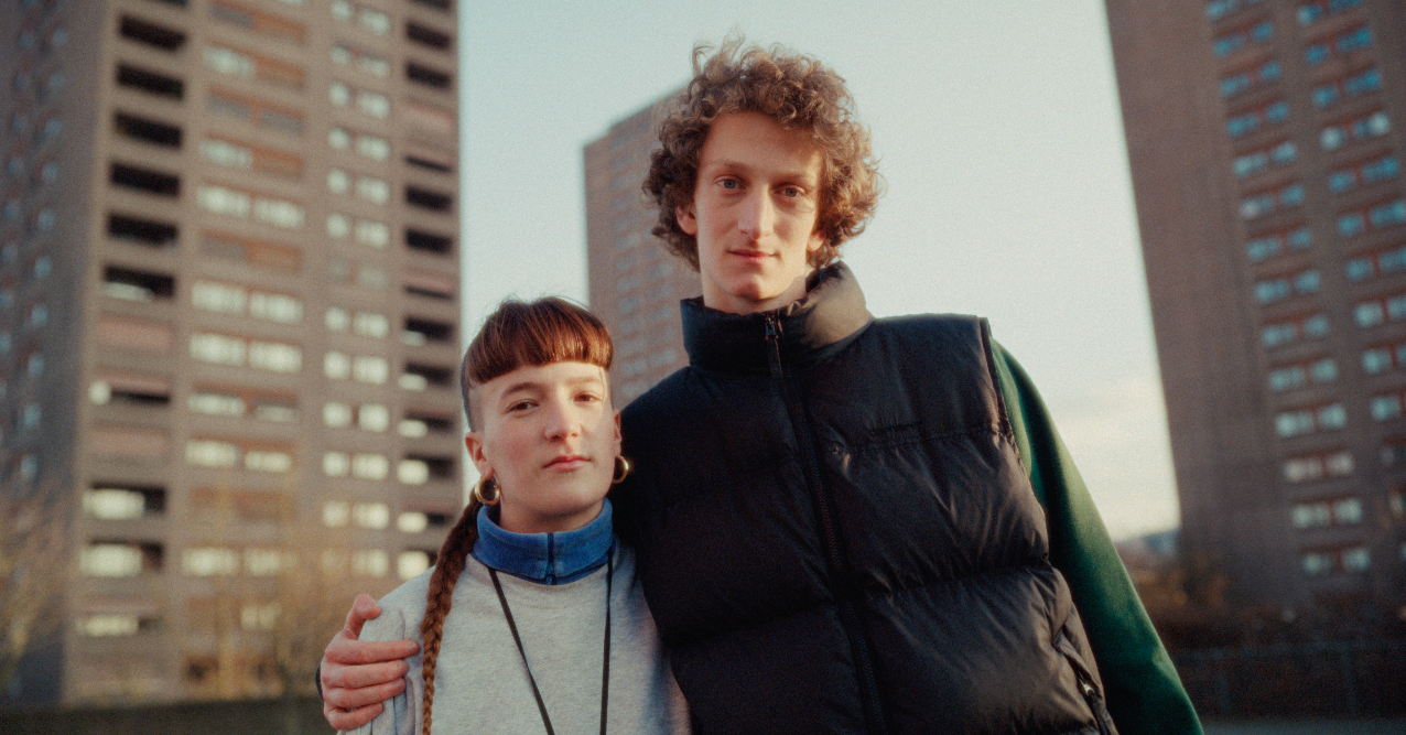A young man and young woman standing close together outdoors in front of tall apartment buildings, with the young man placing his arm around the young woman.