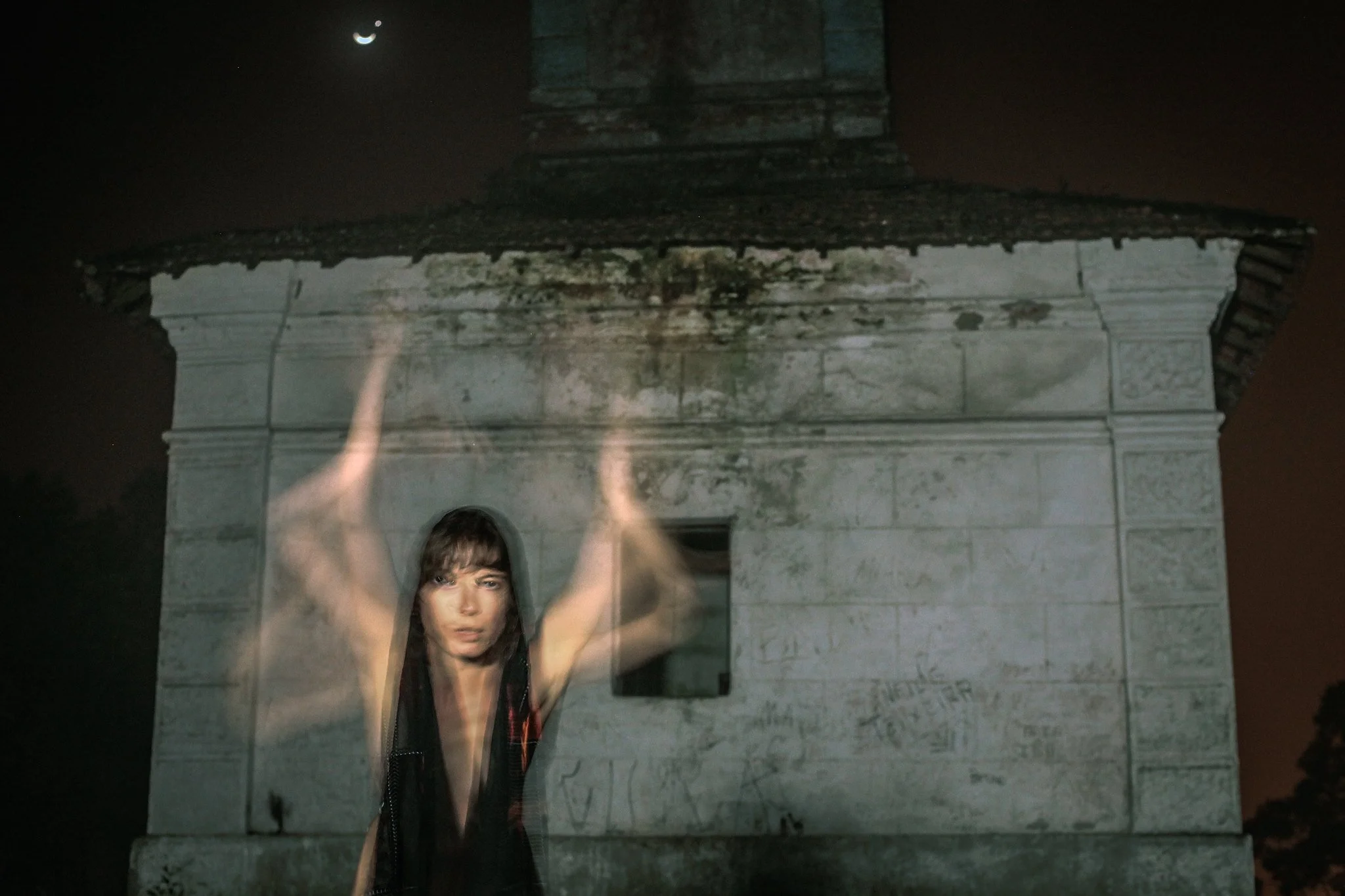 A person with long brown hair and wearing a dark sleeveless top standing behind a glass panel at night, with their arms raised above their head, in front of a historic stone wall with inscriptions, under a night sky with a crescent moon.