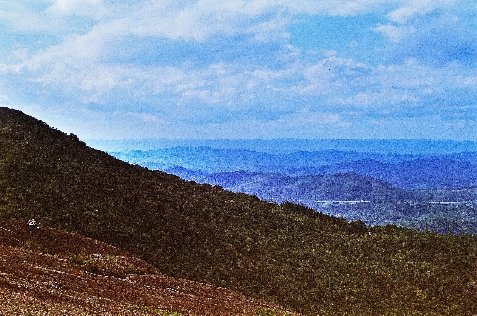 A scenic view of rolling mountains with lush green trees on the slopes, extending into the distance under a partly cloudy sky.