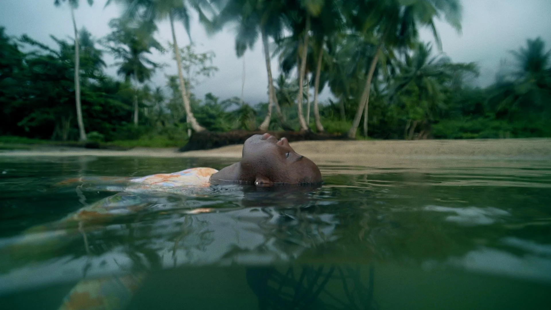 A woman floating in the water with tropical trees and a beach in the background.
