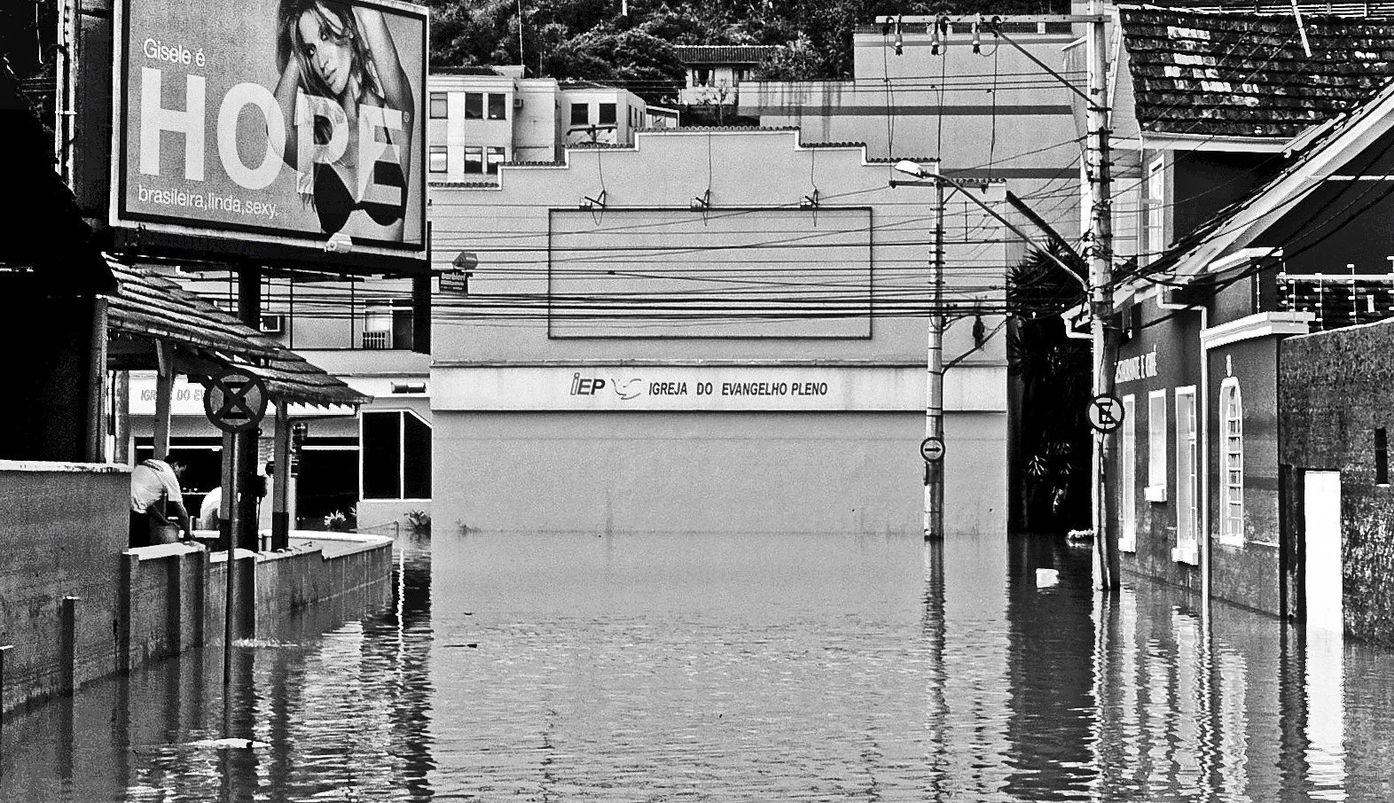 Street flooded with water, submerged sidewalk and street signs, buildings, power lines, and large billboard in the background.