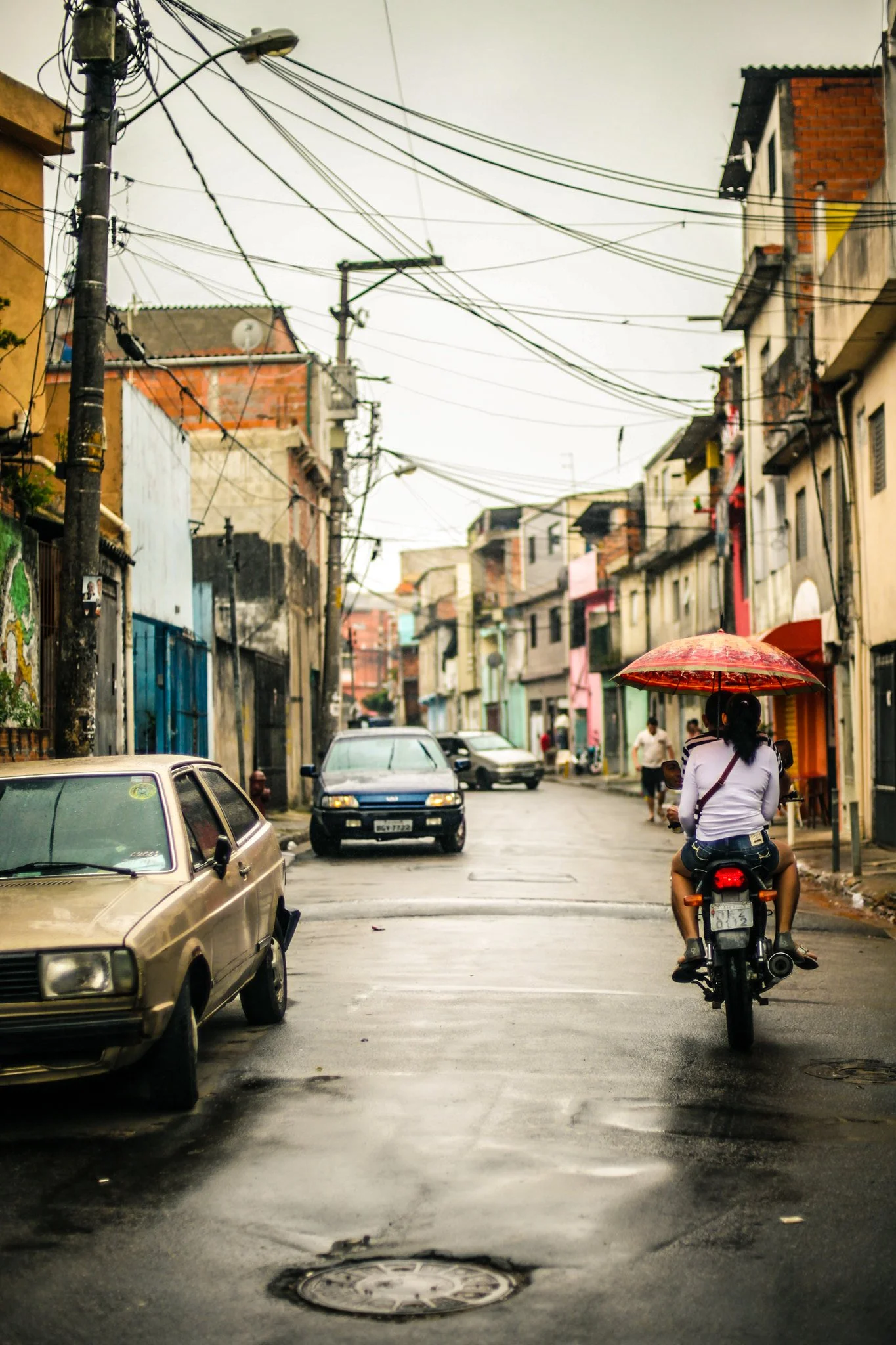 A street scene in an urban neighborhood with parked cars, colorful buildings, and overhead utility wires, featuring a person riding a motorcycle with an umbrella attached to the back in rainy weather.