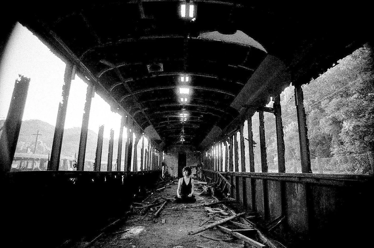 Black and white photo of an abandoned train station platform with debris, broken windows, and a person sitting on the ground in the middle of the platform.