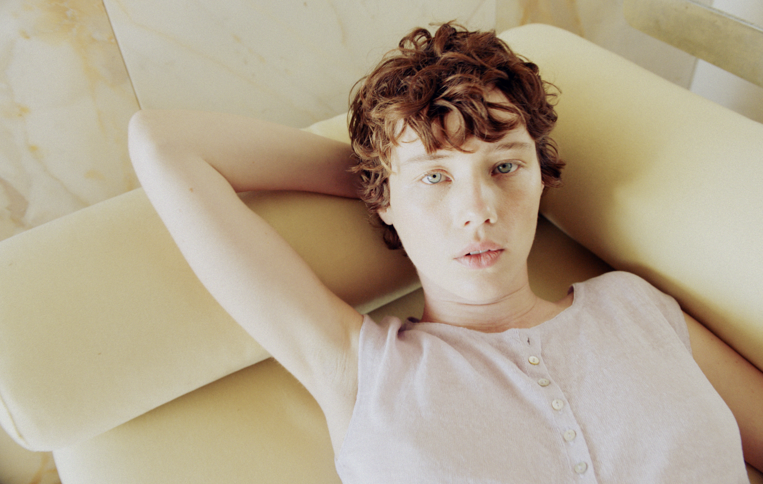 A woman with short curly hair and light eyes is lying on a yellow surface with her hands behind her head, looking directly at the camera.