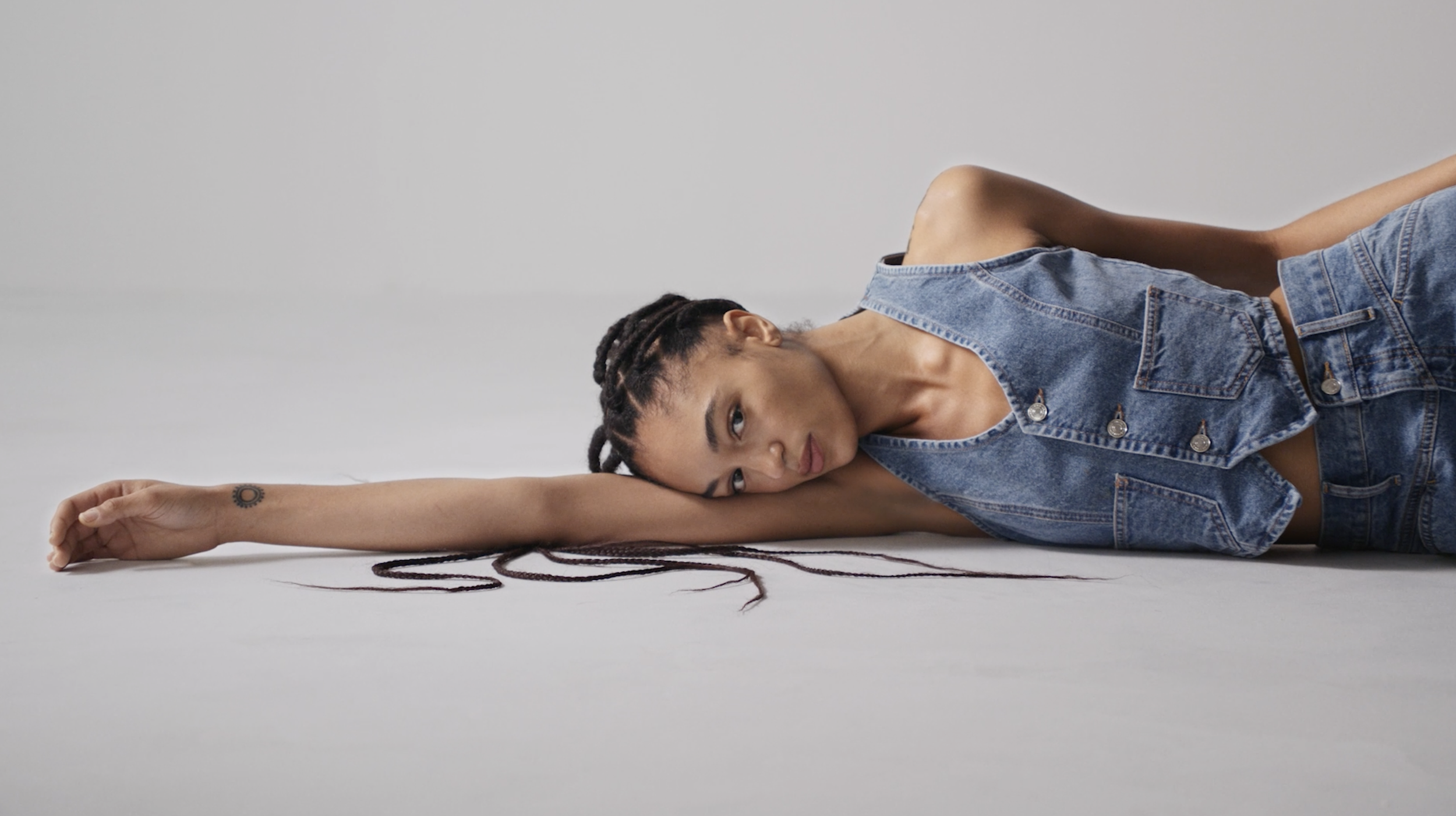 A woman with braids and a tattoo on her wrist laying on the floor in a relaxed pose, stretching her arm with her head resting on her arm, wearing a denim vest and jeans, against a plain light background.