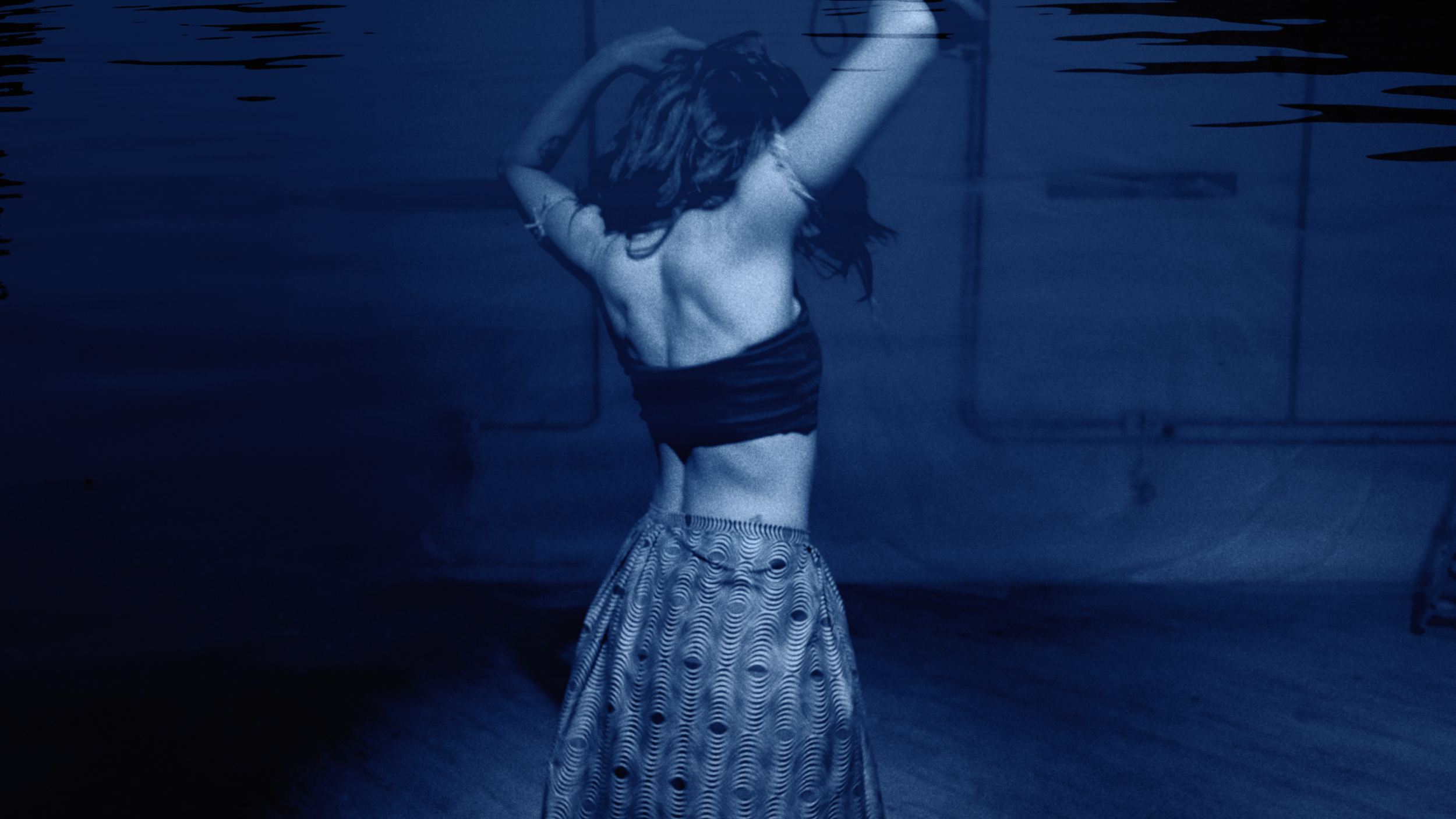 A person with long hair wearing a striped skirt and black shorts under water with their hand behind head.