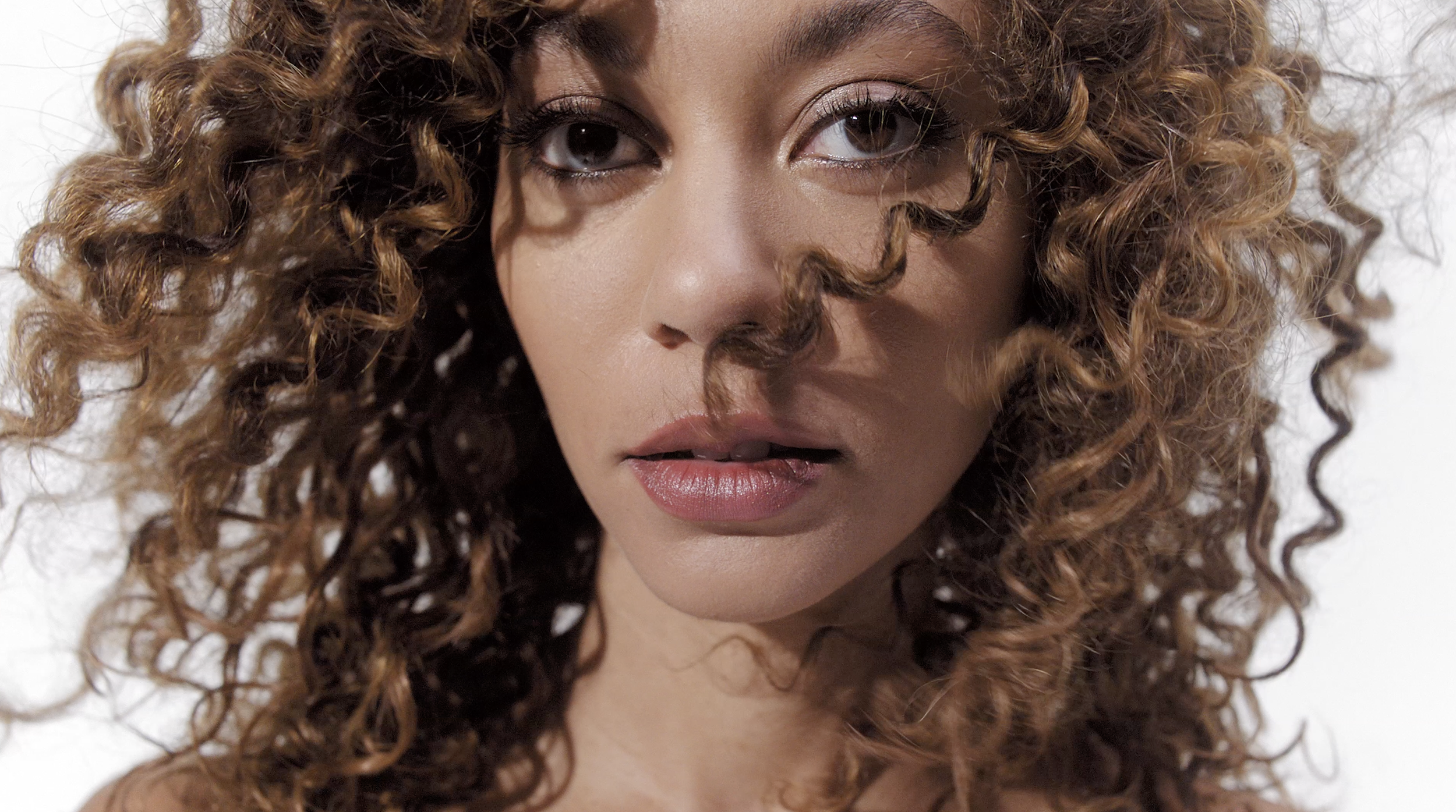 Close-up of a woman with curly brown hair and makeup, looking directly at the camera.