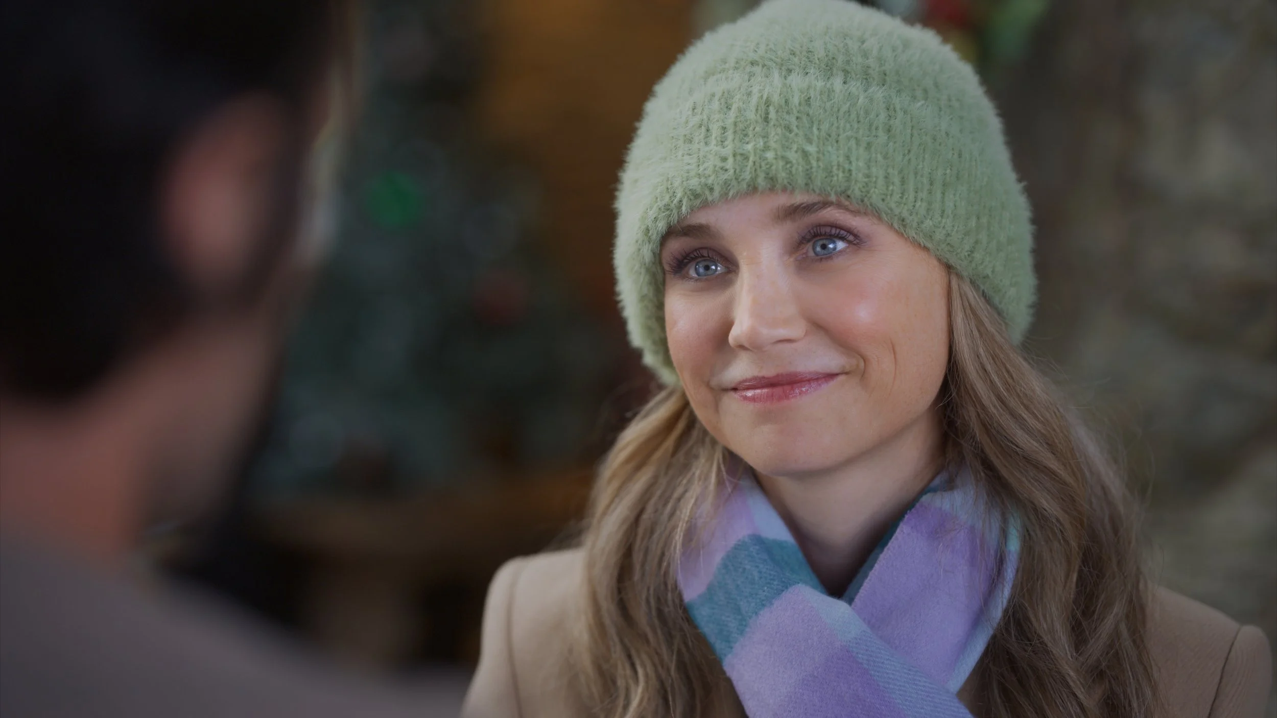 A woman with blue eyes, long curly hair, wearing a light green knit beanie and a purple scarf, smiles at someone