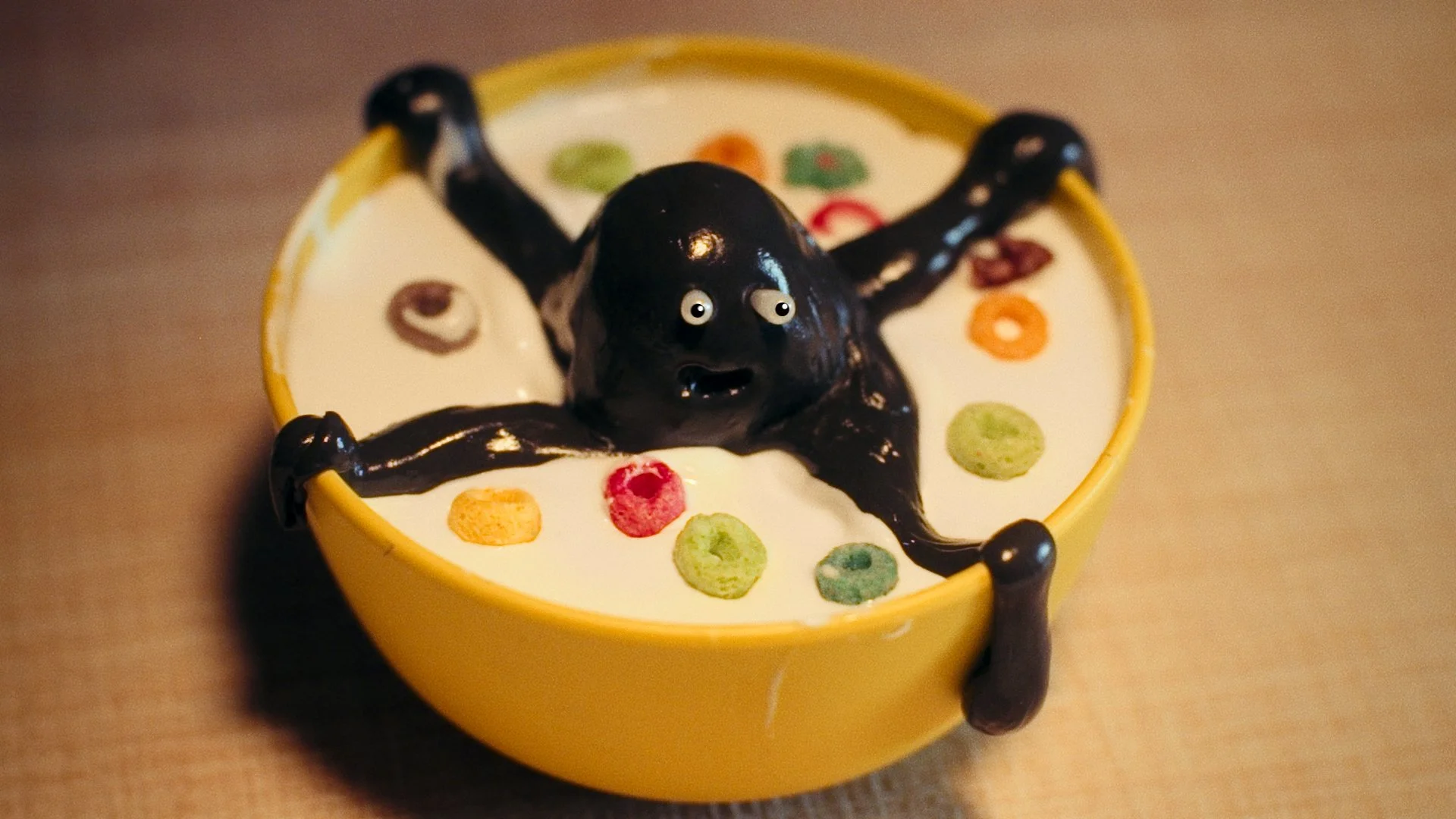 A small yellow bowl filled with white liquid, decorated with colorful cereal pieces, has a black octopus-shaped figure with detailed eyes and a mouth submerged in the liquid.
