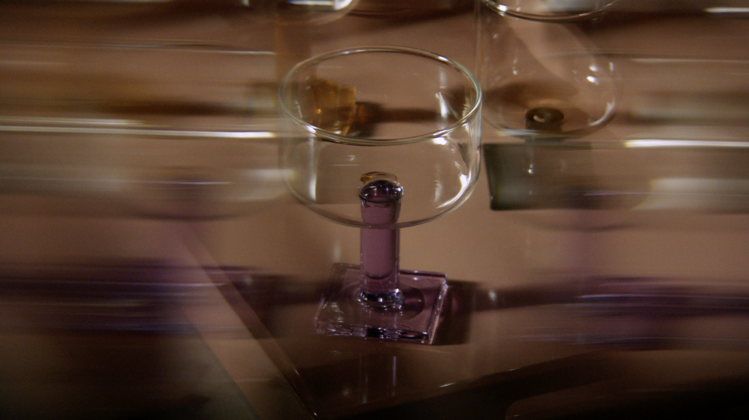 A clear cocktail glass with a pink stem on a reflective surface, reflecting the glass and surrounding objects.