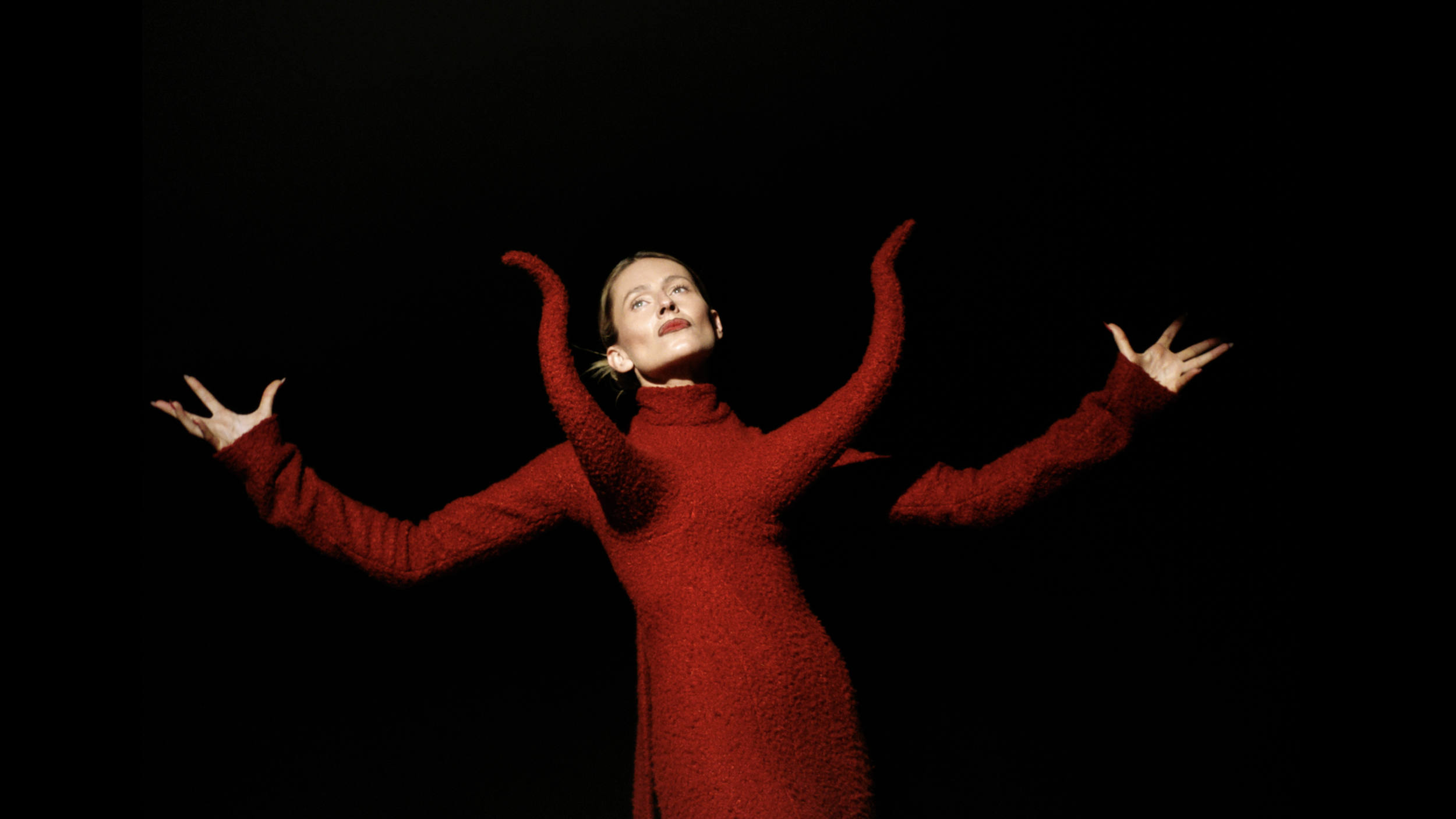 A woman with short hair and red lipstick wearing a red fuzzy turtleneck sweater with arms outstretched against a black background.