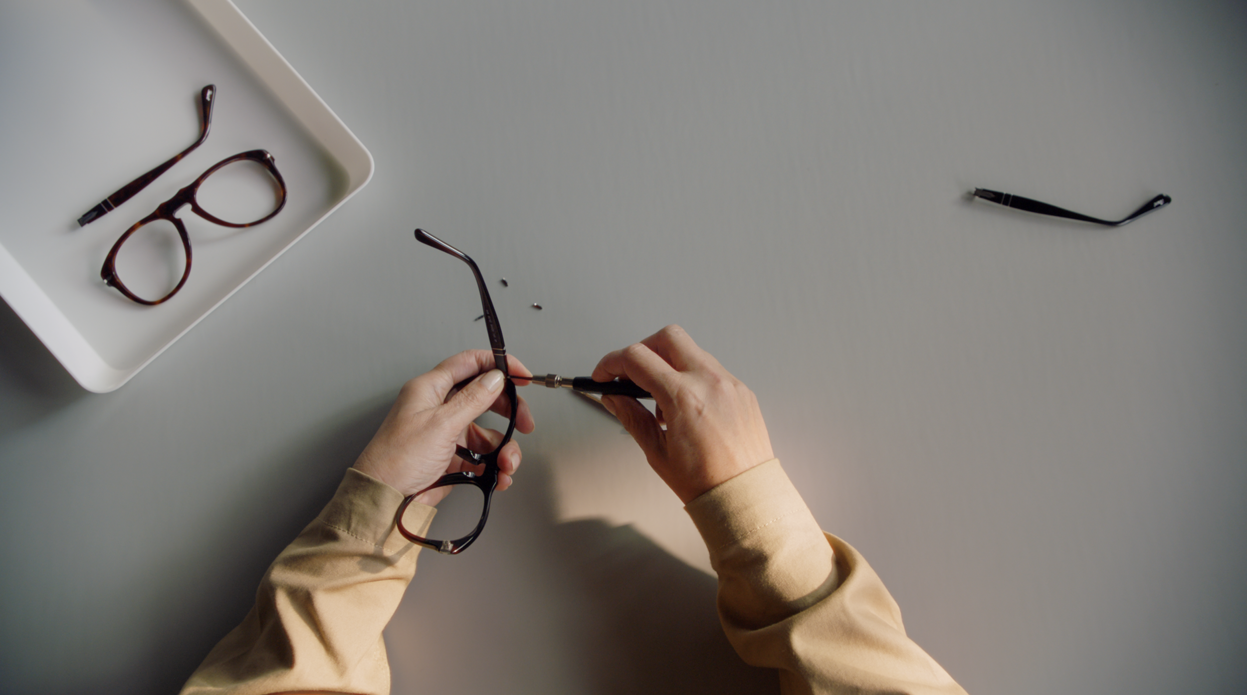 Person holding a pair of glasses and a screwdriver, repairing or assembling glasses on a gray table. A white tray with additional glasses rests nearby.