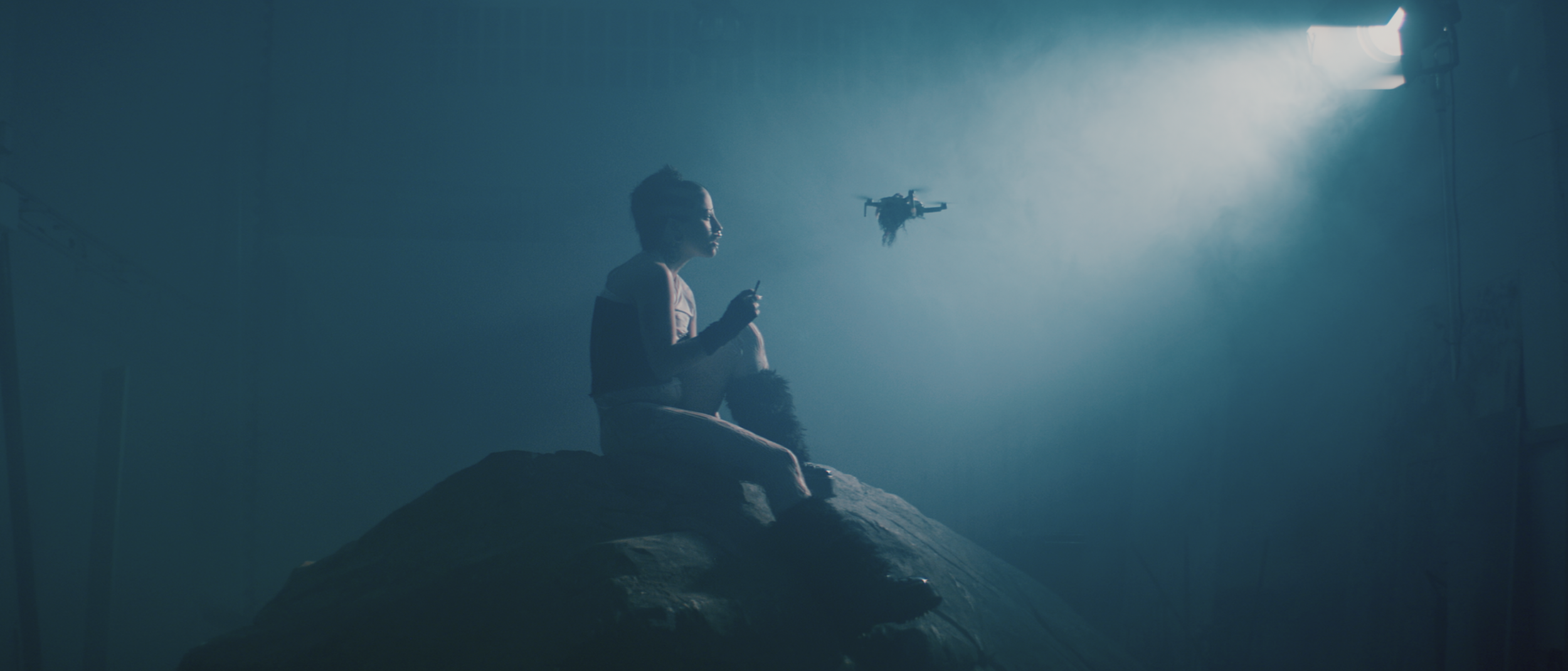 A woman sitting on a rock in a dimly lit room with blue tint, holding a remote control, with a drone hovering nearby and a soft light illuminating the scene from the upper right corner.